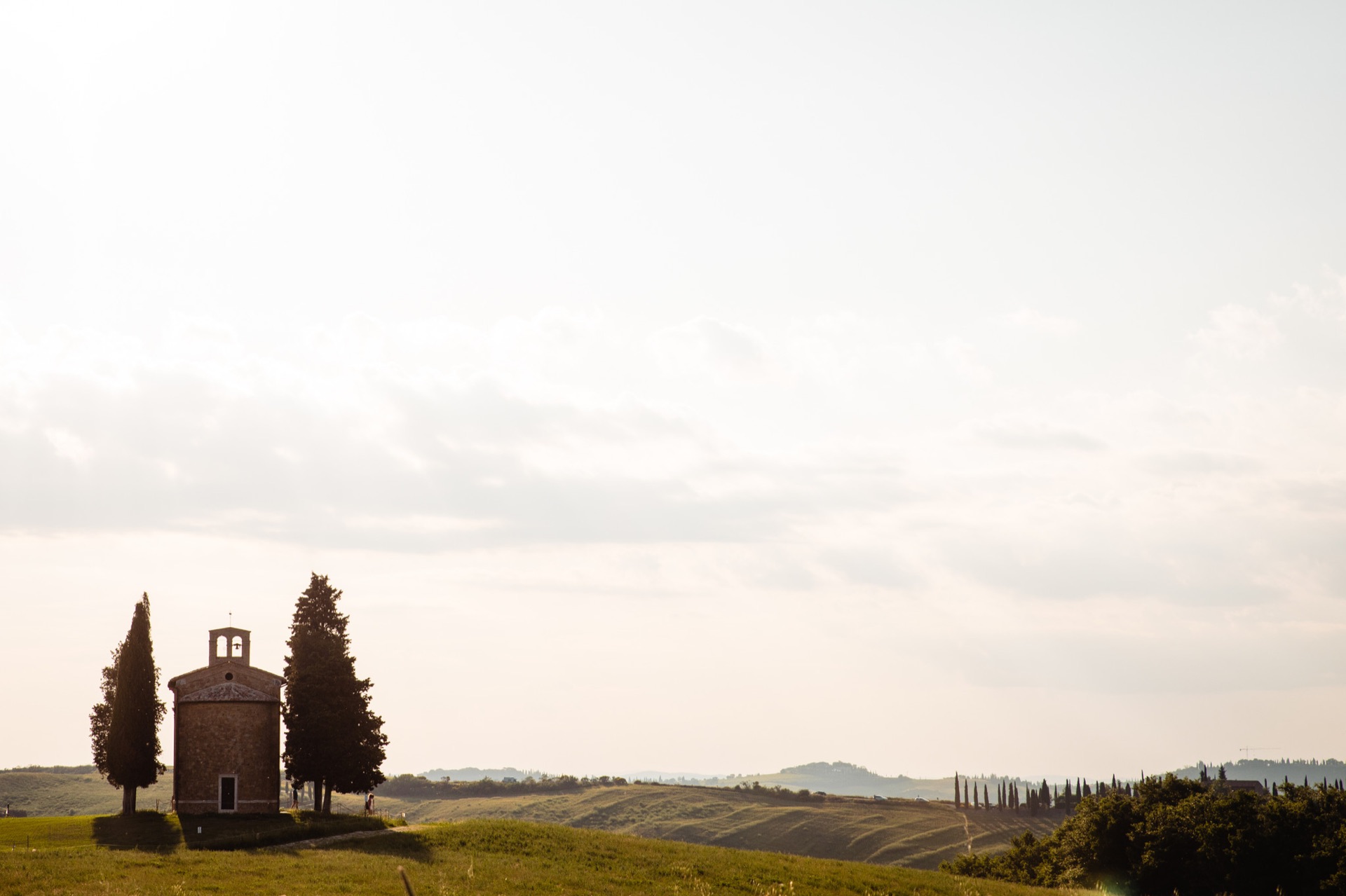 A small chapel among cypress trees and hills beneath a luminous Tuscan sky