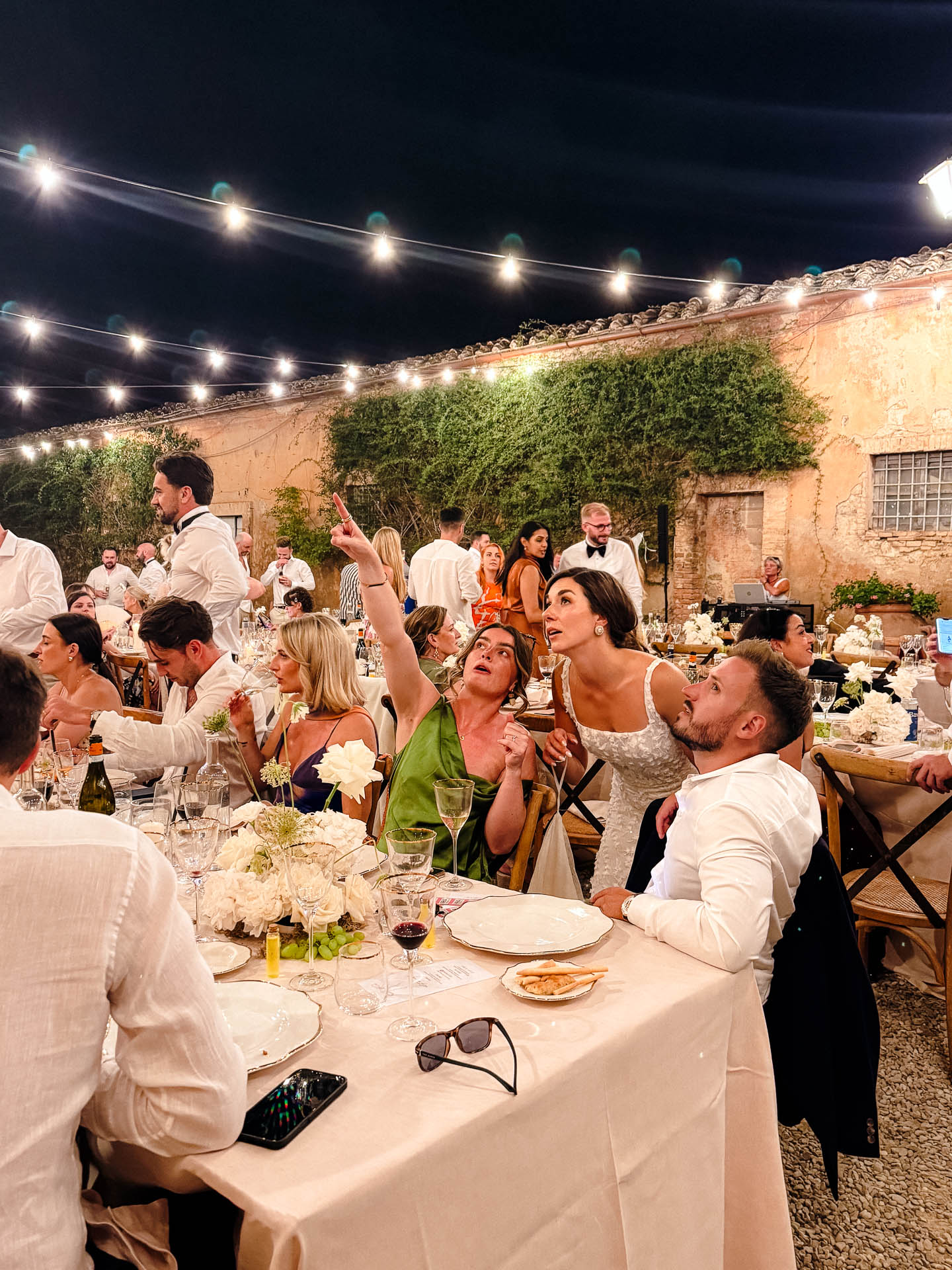 Outdoor night wedding reception with string lights, guests seated at long tables, floral centerpieces, ivy-covered walls in the background.