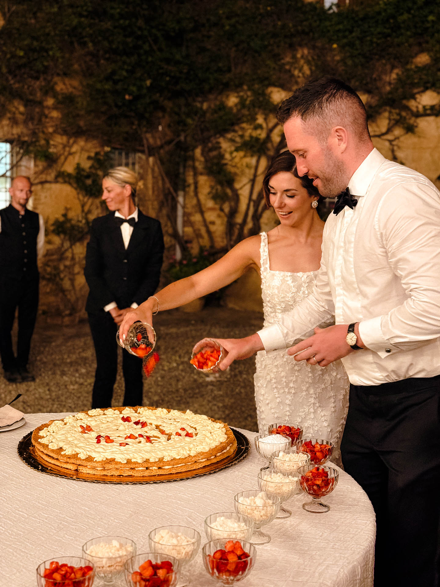 Bride and groom in formal attire slice a large cream-topped cake at an outdoor celebration, with small bowls of fruit nearby and guests in suits watching in the background.