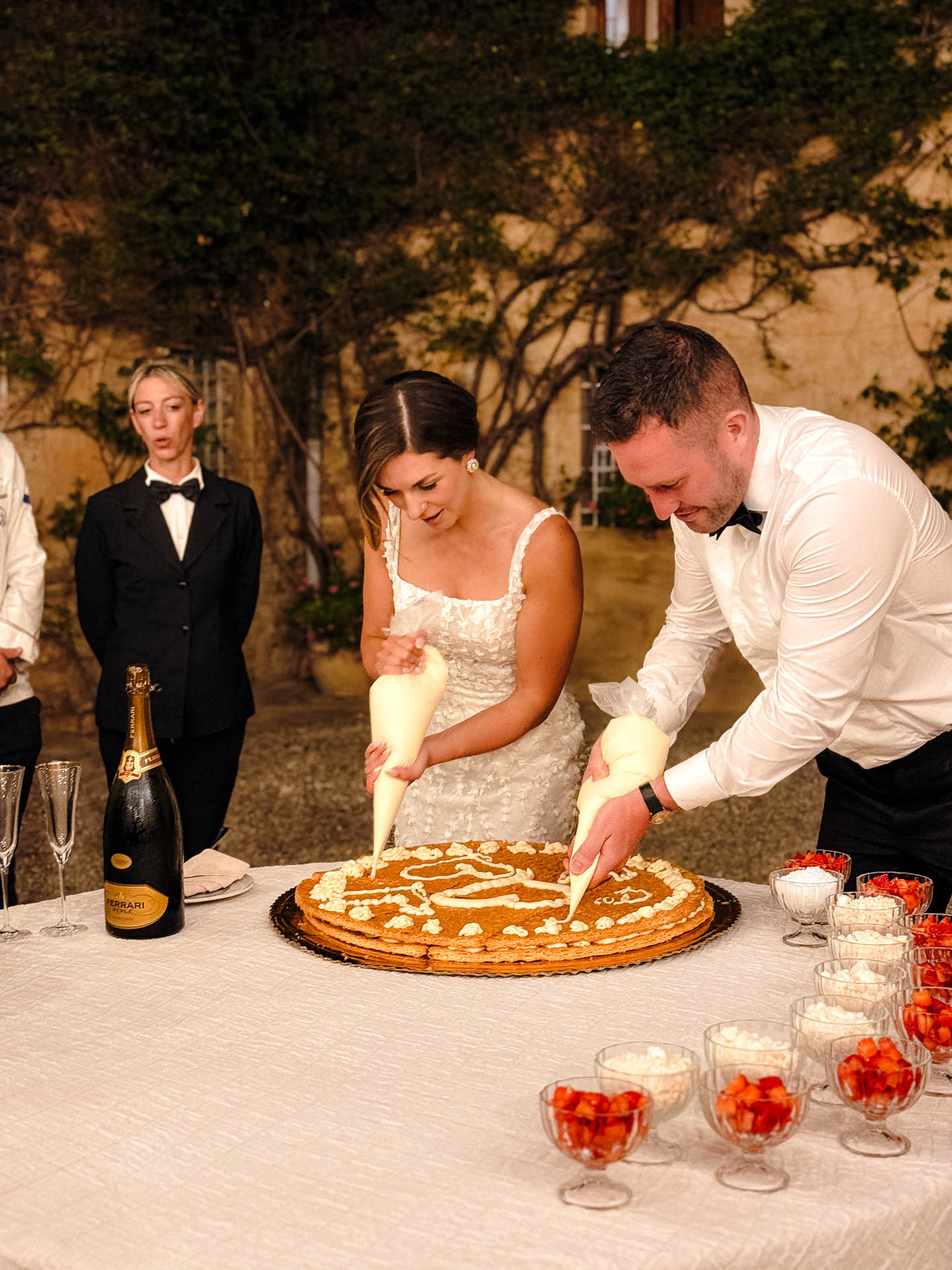 Bride in a white dress and a man in a white shirt slice a large round cake at an outdoor wedding reception.