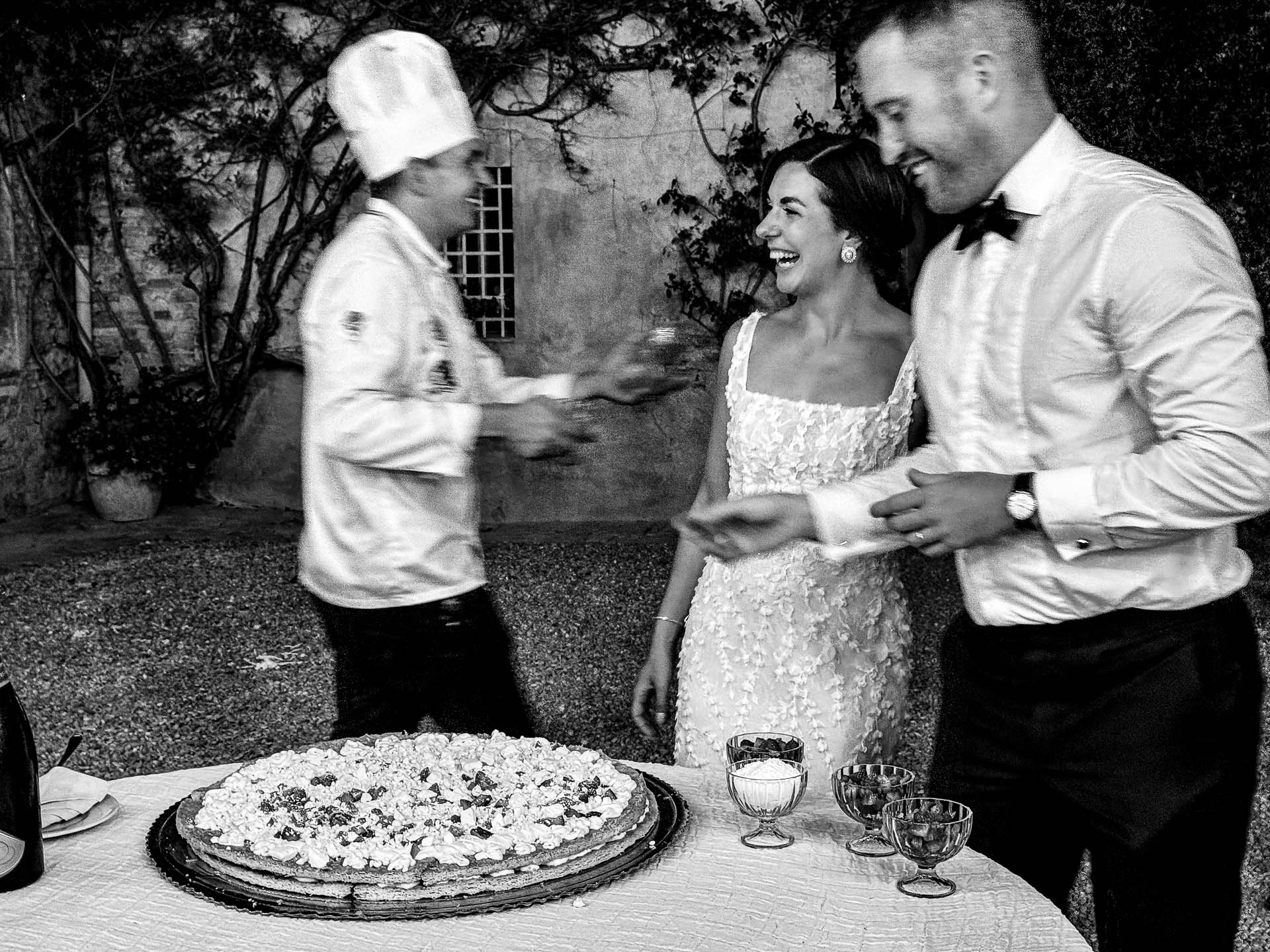 Bride in a lace dress smiles as a server approaches with plates beside a large, frosted wedding cake on an outdoor table.