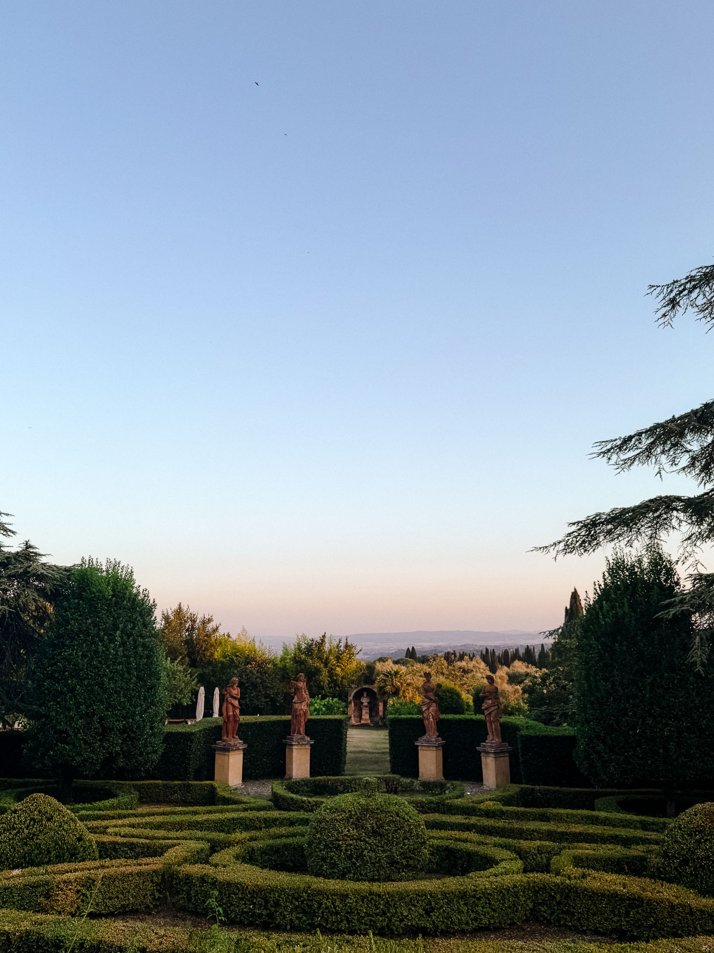 Formal garden with statues on pedestals along a neatly trimmed hedge path, framed by tall trees and a distant horizon under a clear sky.