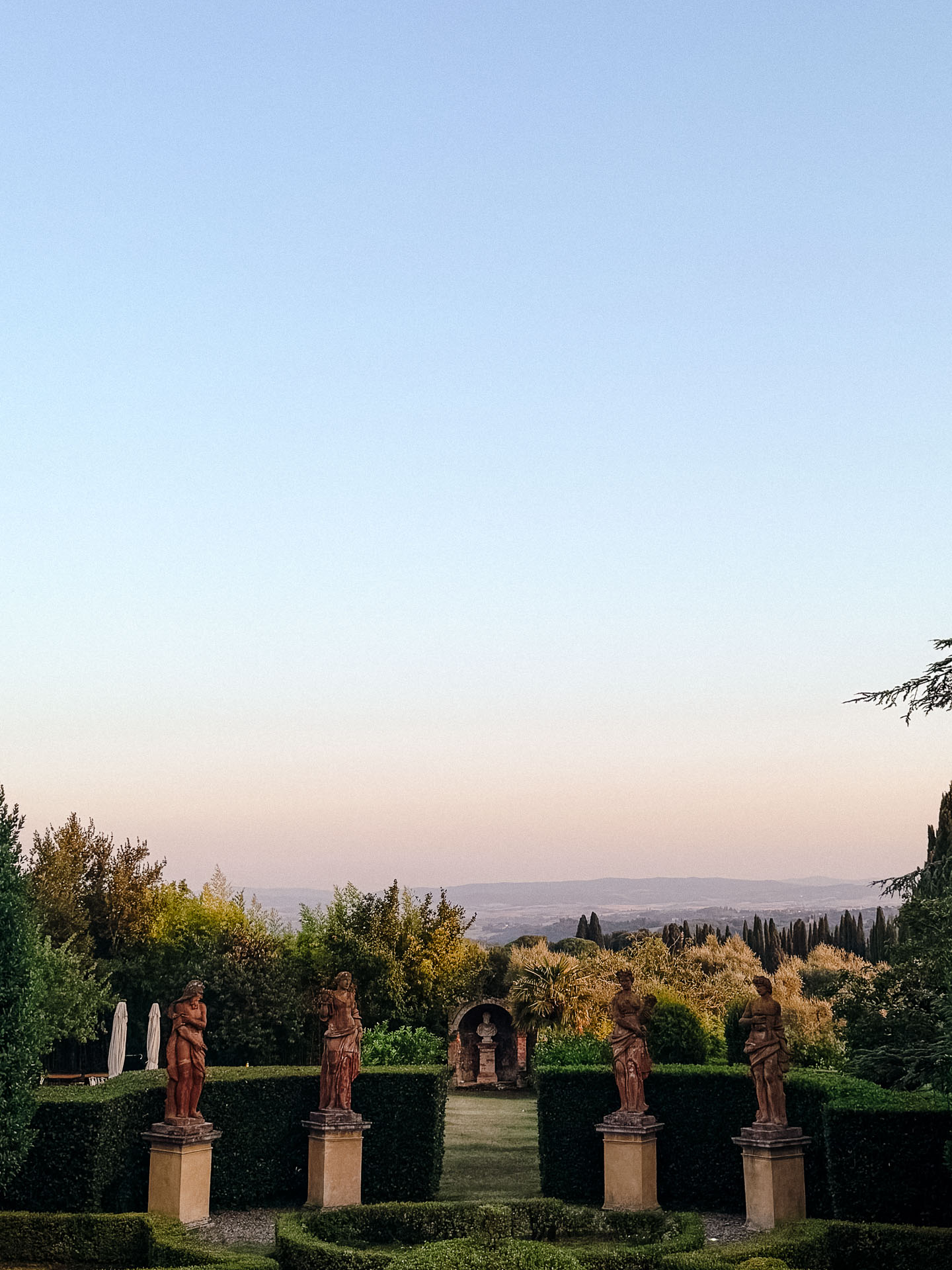 Formal garden with a row of classical statues on pedestals flanking trimmed hedges, under a sunset sky.