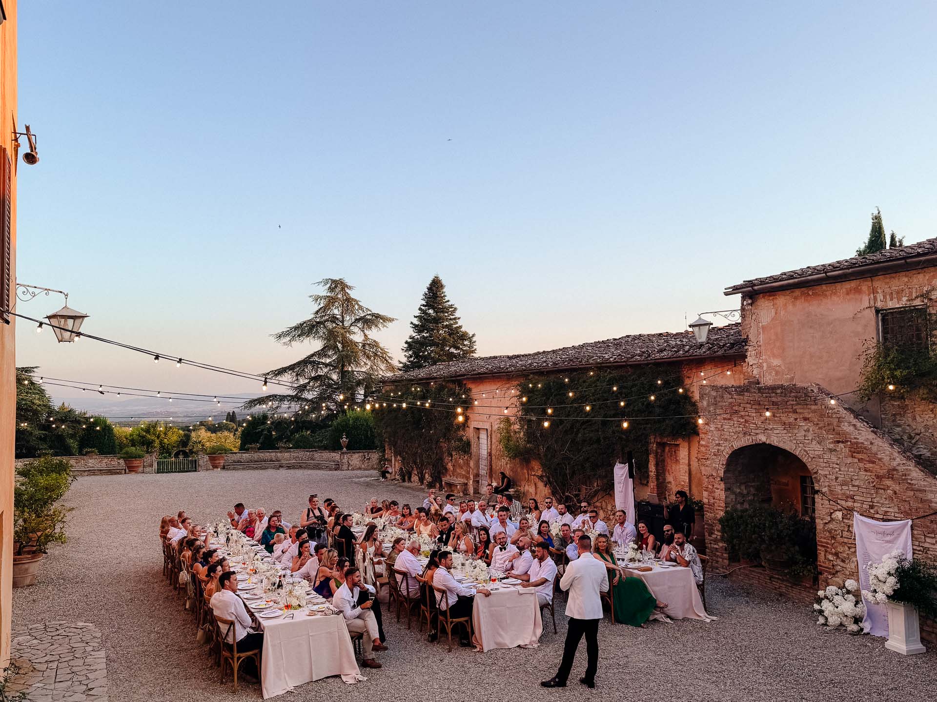 Outdoor wedding reception in a sunlit courtyard with long banquet tables and string lights above.