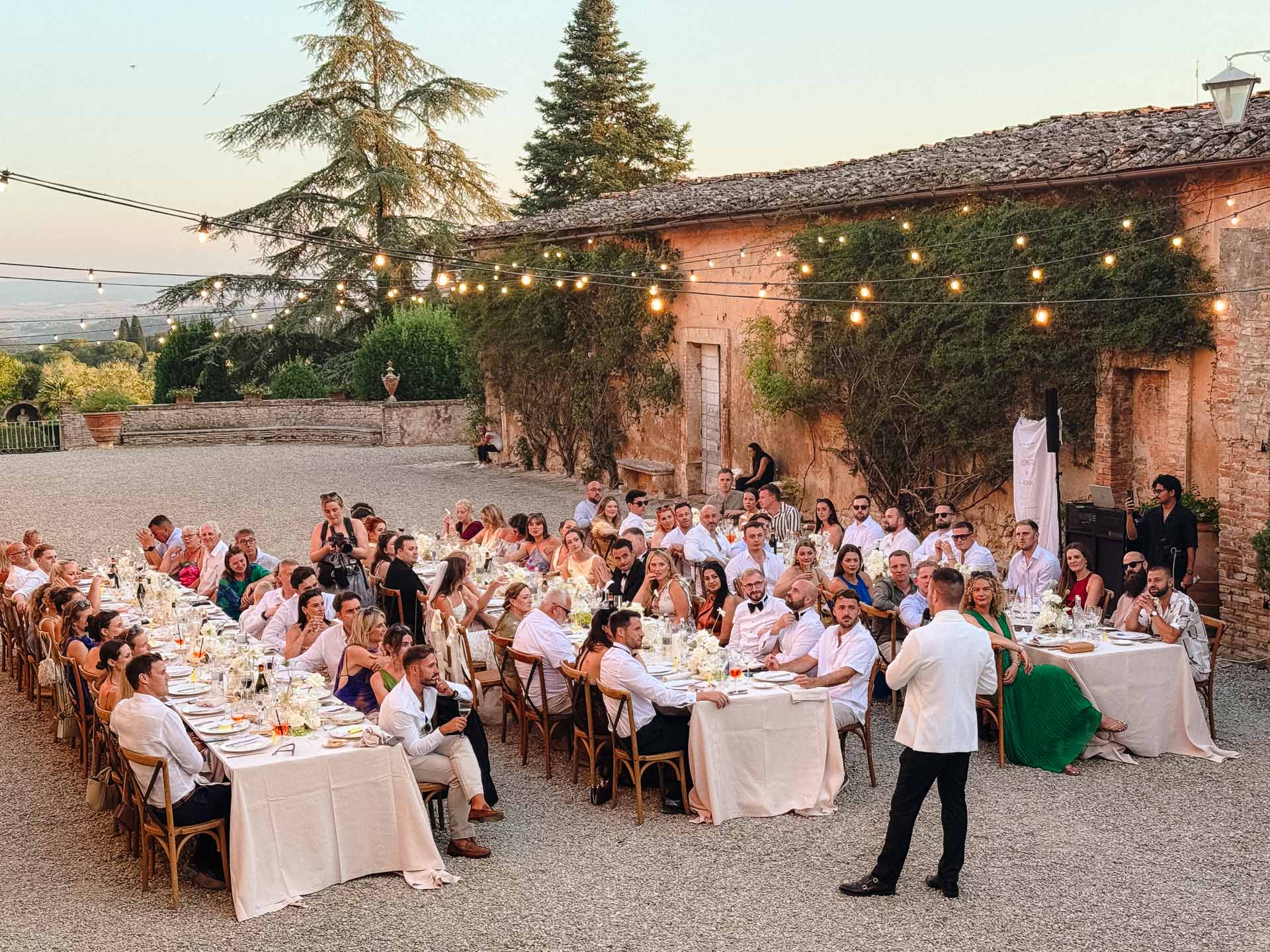 Outdoor wedding reception with long white-clothed tables, guests seated, and string lights overhead in a rustic courtyard