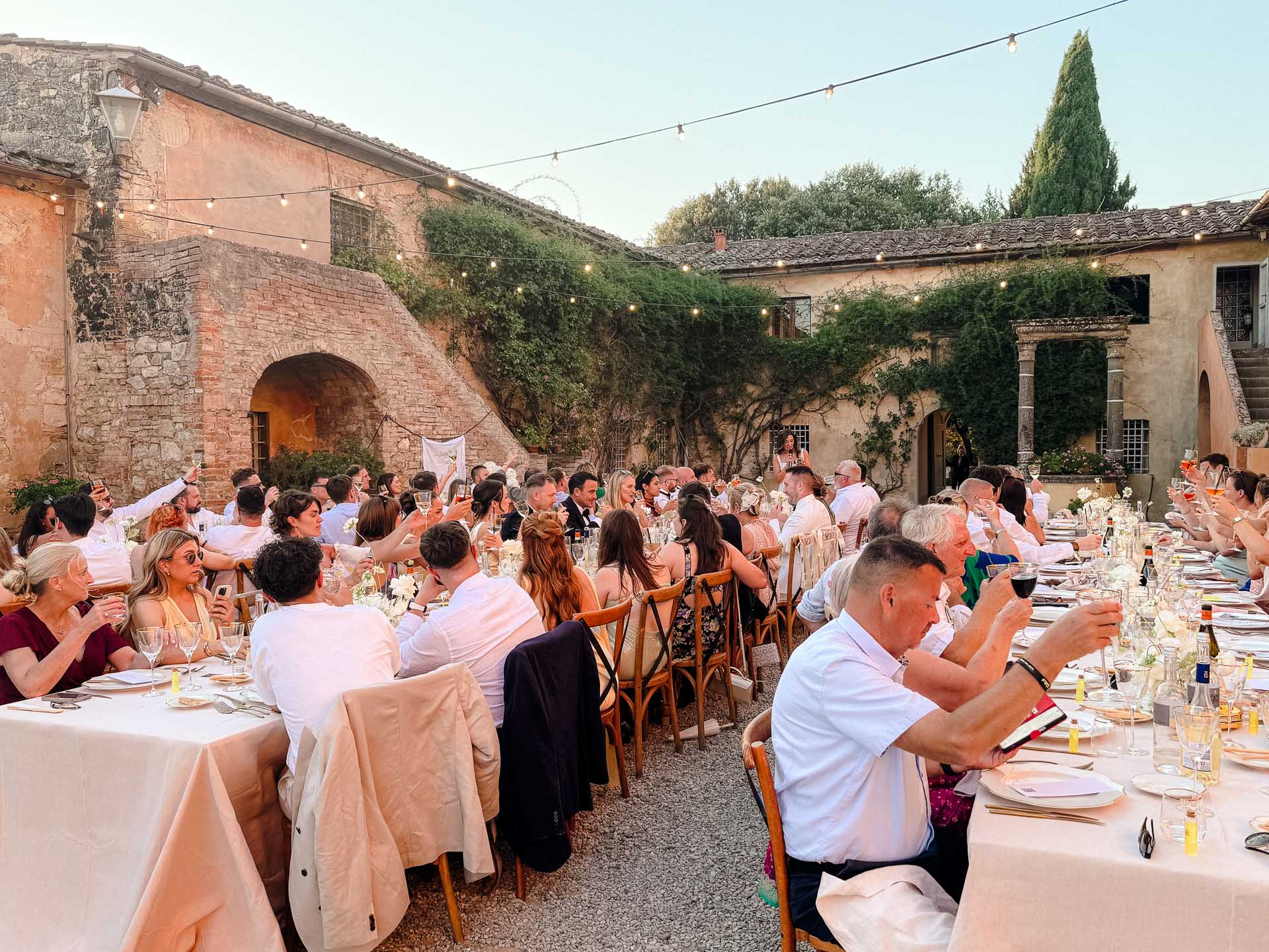 Outdoor wedding reception in a stone courtyard with long tables and string lights, guests toasting drinks under a warm evening sky.