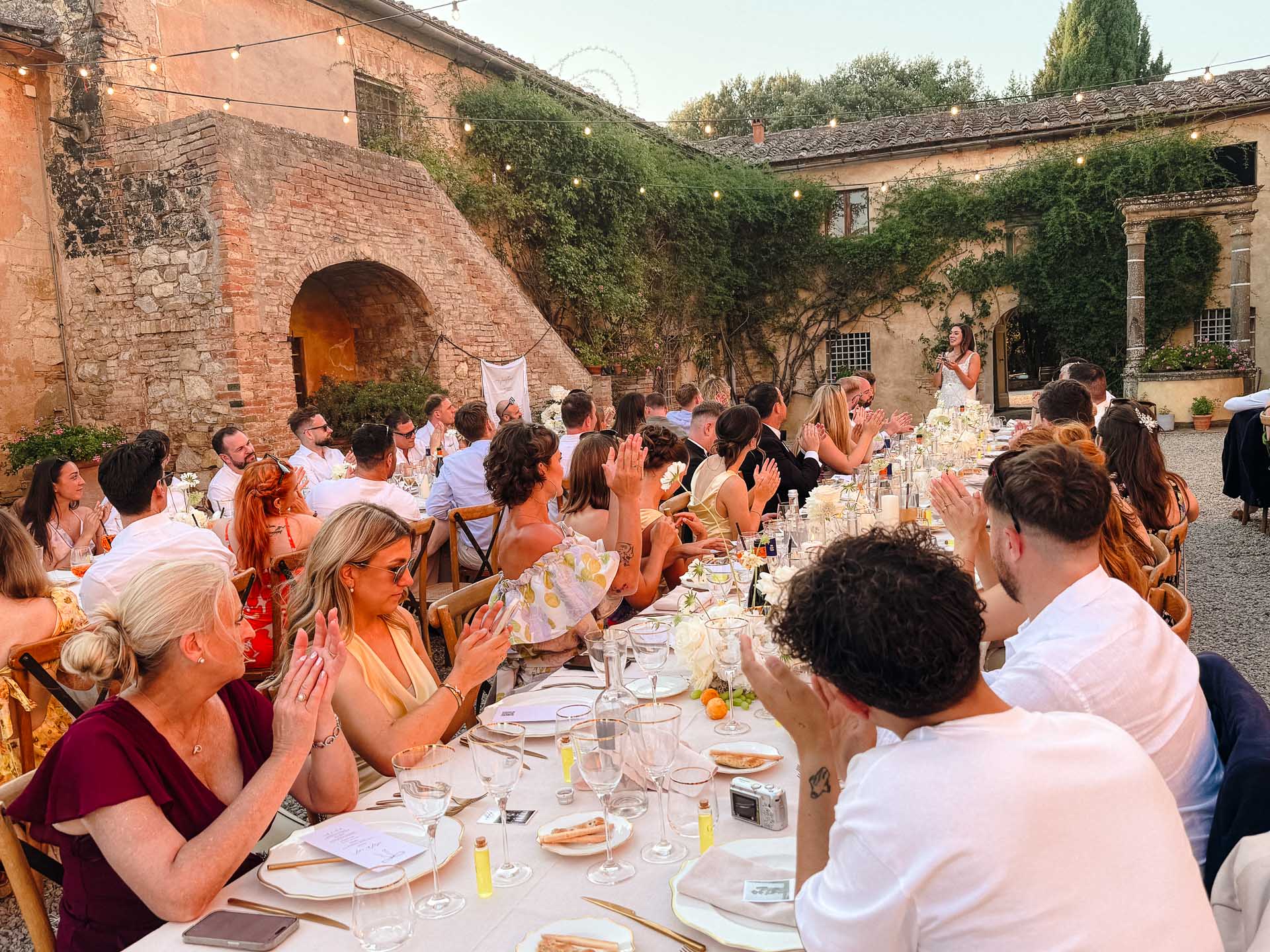 Outdoor wedding reception with a long table of guests clapping as a speaker stands at the end.