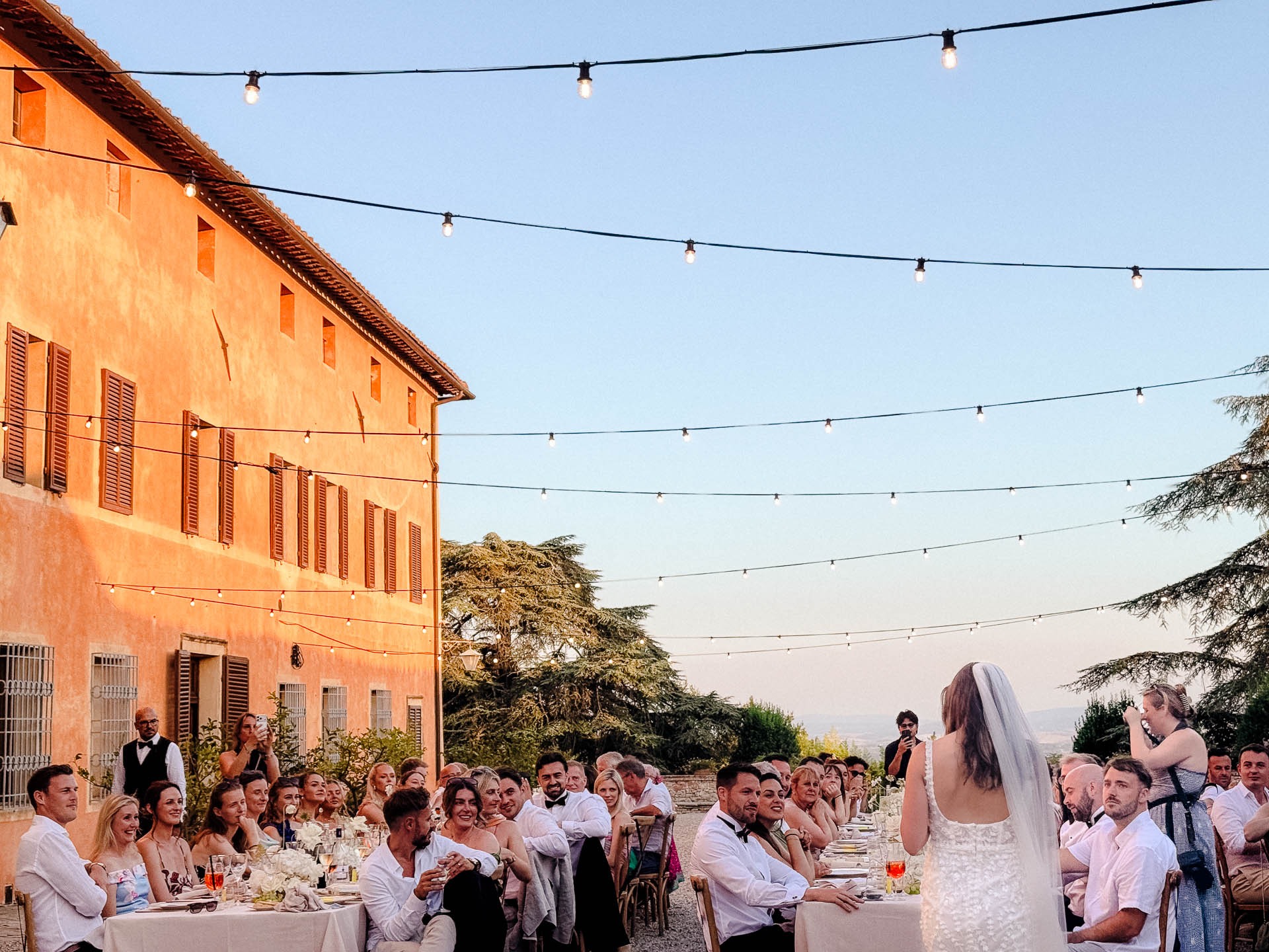 Outdoor wedding reception with guests seated at long tables under string lights, bride in white lace dress addressing the crowd.