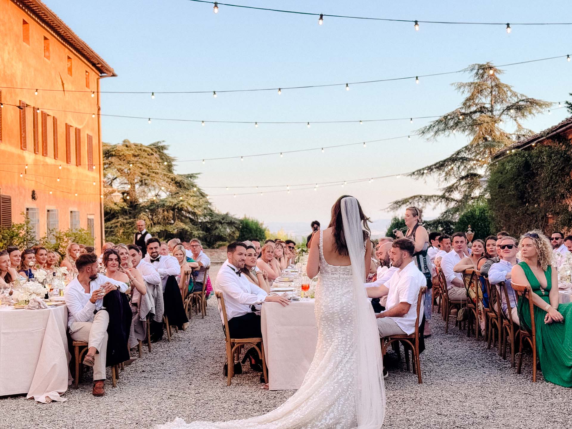Bride in a lace wedding gown with long train and veil speaks to guests at an outdoor reception under string lights.