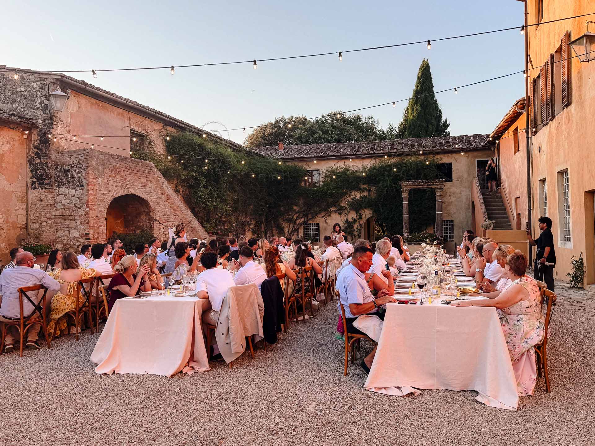 Outdoor wedding reception in a sunlit stone courtyard with long banquet tables and string lights overhead, guests dining together.