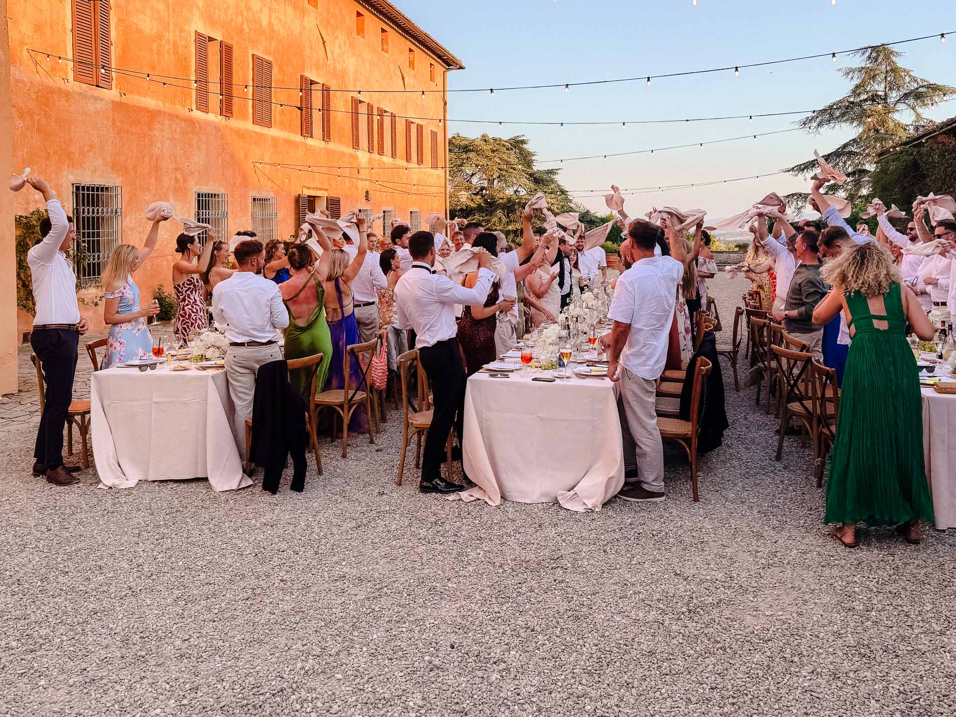 Outdoor wedding reception with guests standing and waving napkins around long, white-clothed tables under string lights atop an orange building.