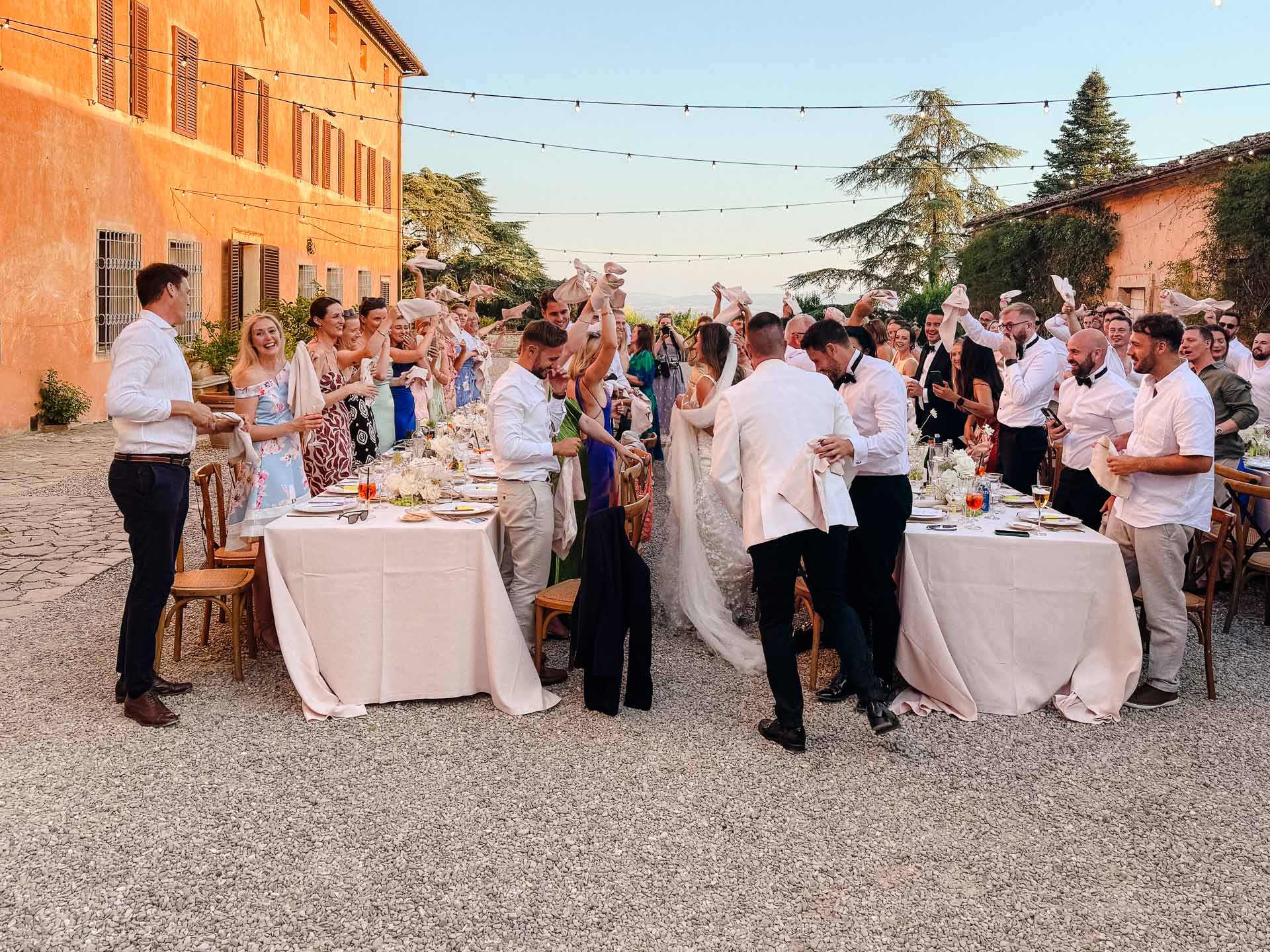 Outdoor wedding reception with guests raising napkins in celebration along long banquet table under string lights.