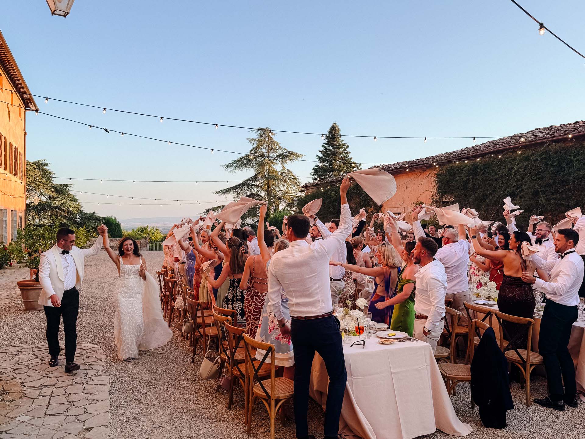Bride and groom enter an outdoor wedding as guests cheer and toss napkins under string lights. The sunlit courtyard is lined with chairs and tables filled with guests in formal attire.",