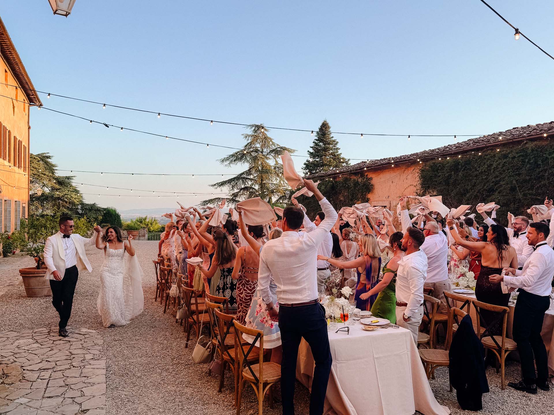 Bride and groom walk through an outdoor wedding reception as guests cheer and toss napkins in the air.