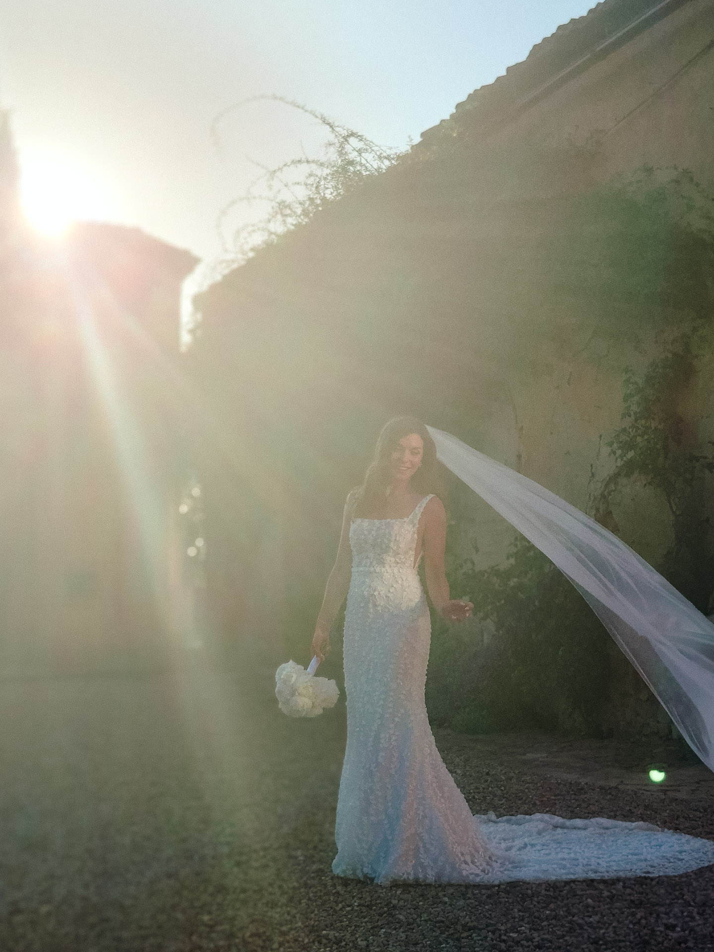 Bride in a lace wedding gown holding a bouquet, veil trailing behind, walking on a gravel path with a sun flare in a sunny outdoor setting.