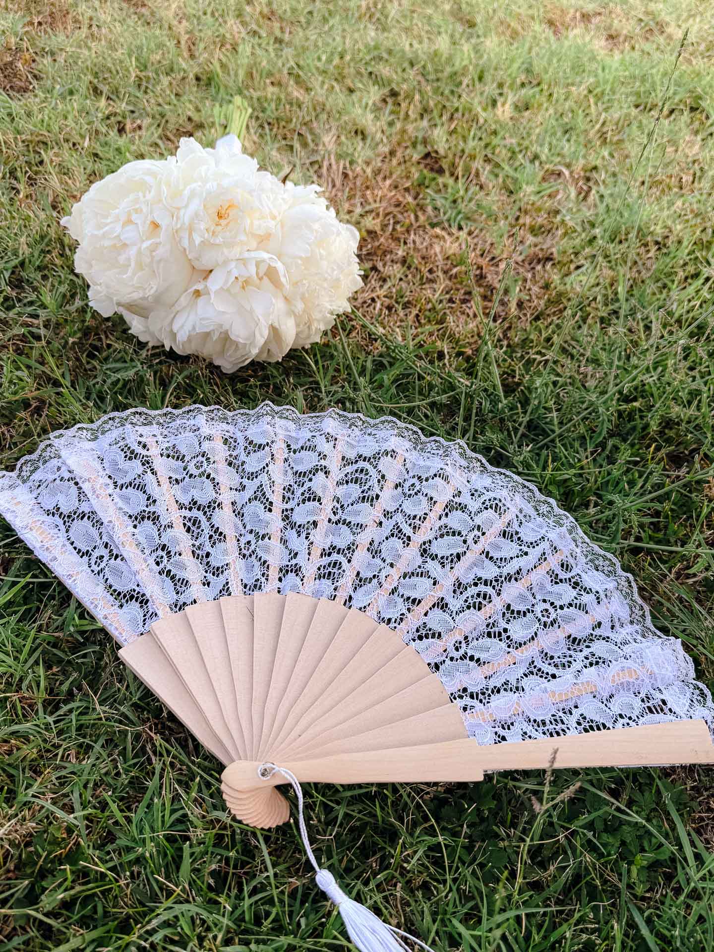 White peony on green grass beside a white lace hand fan, outdoors on a sunny day.