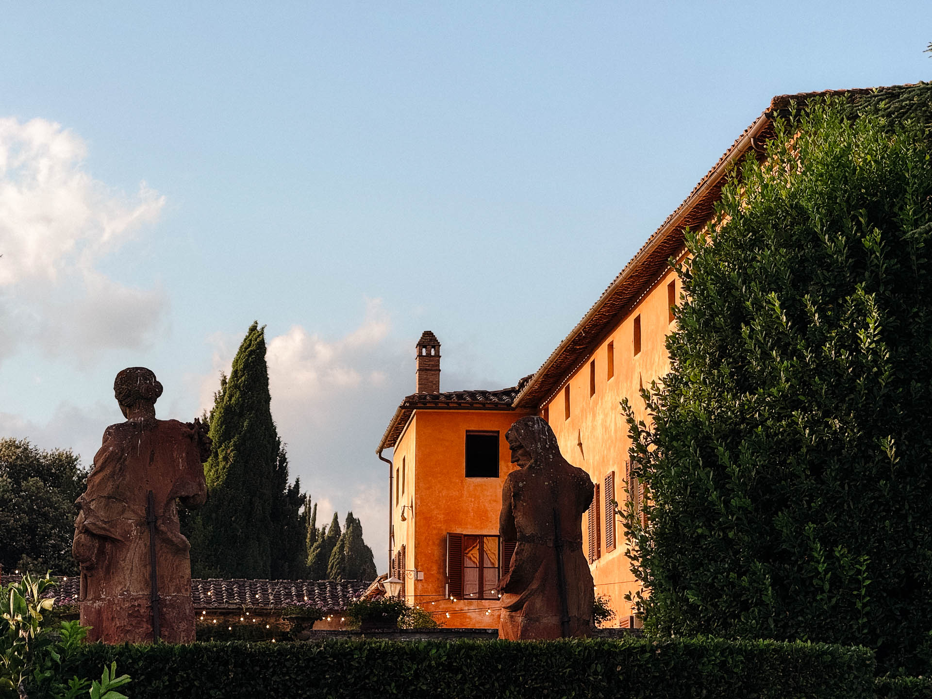 Orange stucco villa with brown shutters, two stone statues in a formal garden and string lights overhead.