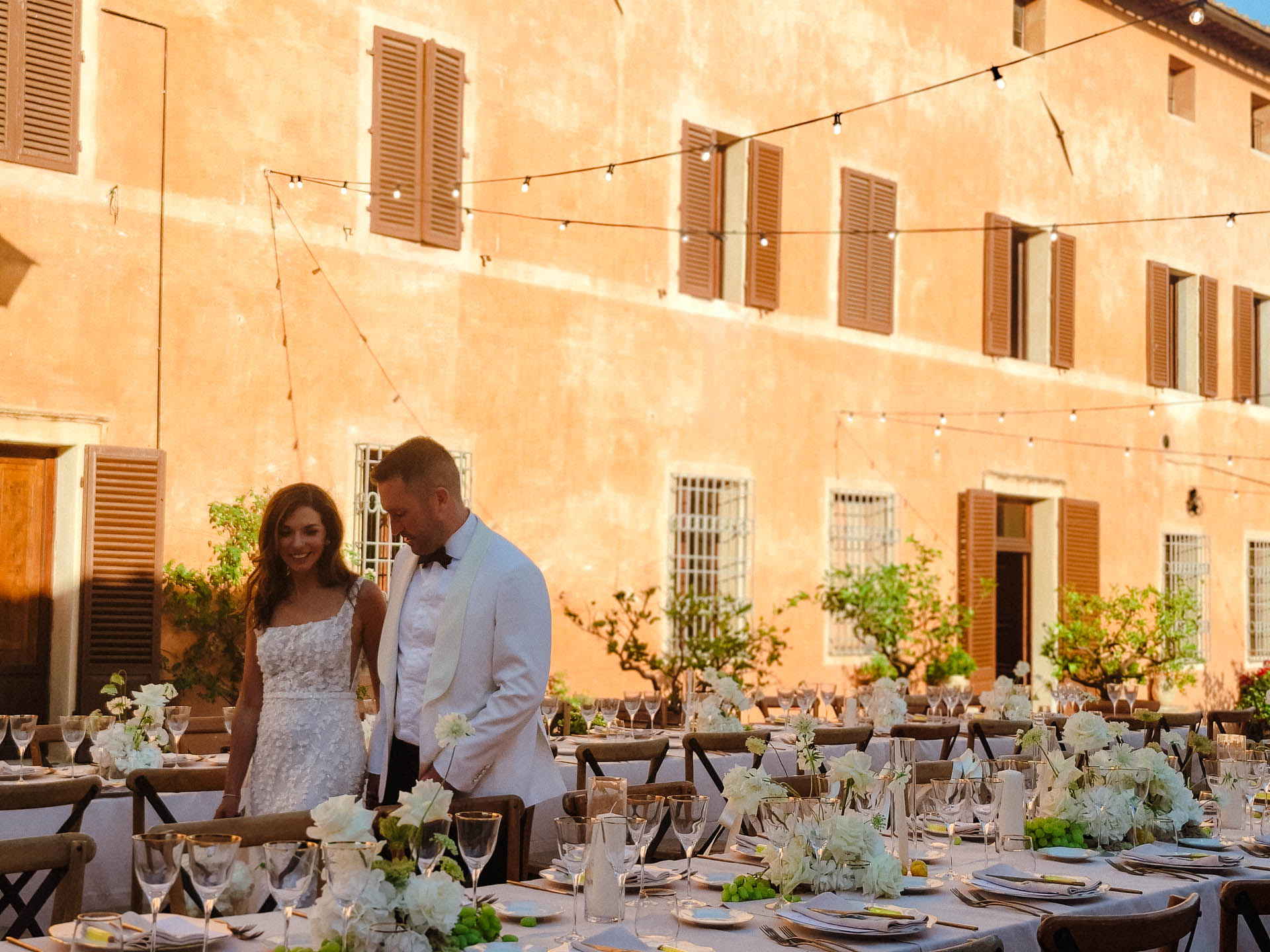 Bride and groom walk past a long, decorated outdoor reception table in a warm peach courtyard with string lights overhead.