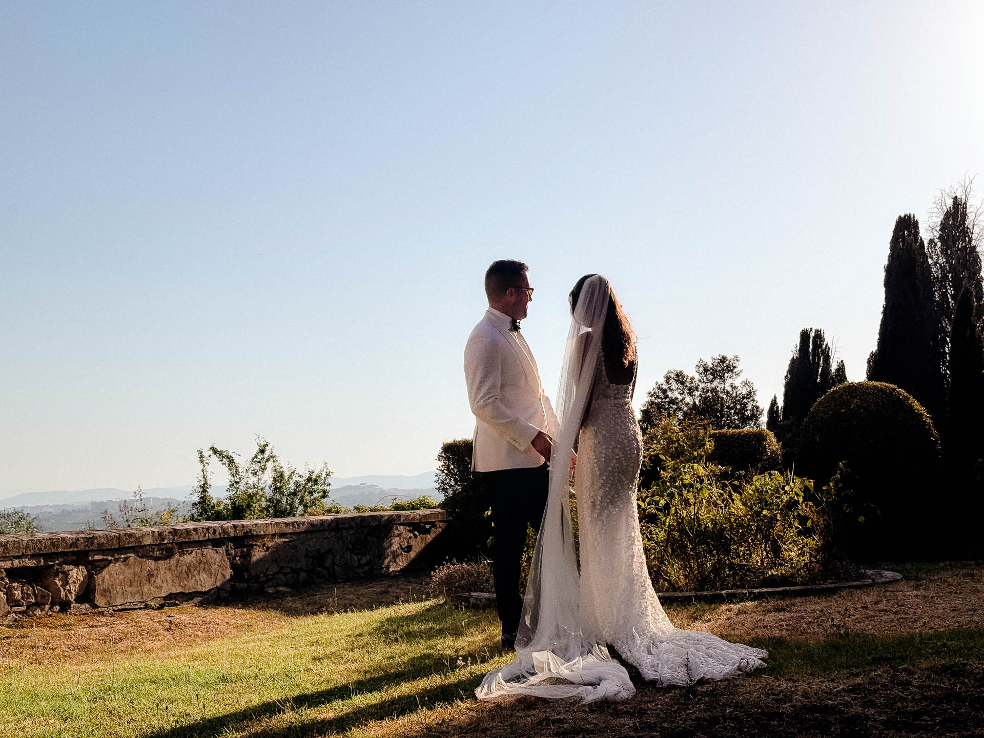Bride and groom in wedding attire stand outdoors beside a stone wall with a clear blue sky behind them.