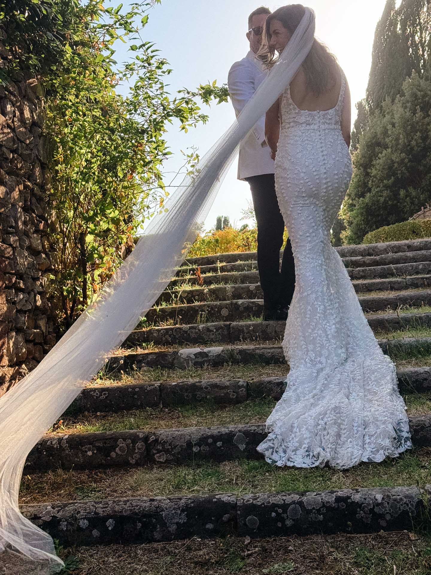 Bride in a white lace wedding gown with a long veil standing on stone steps outdoors, with a man in a white shirt beside her and greenery around.