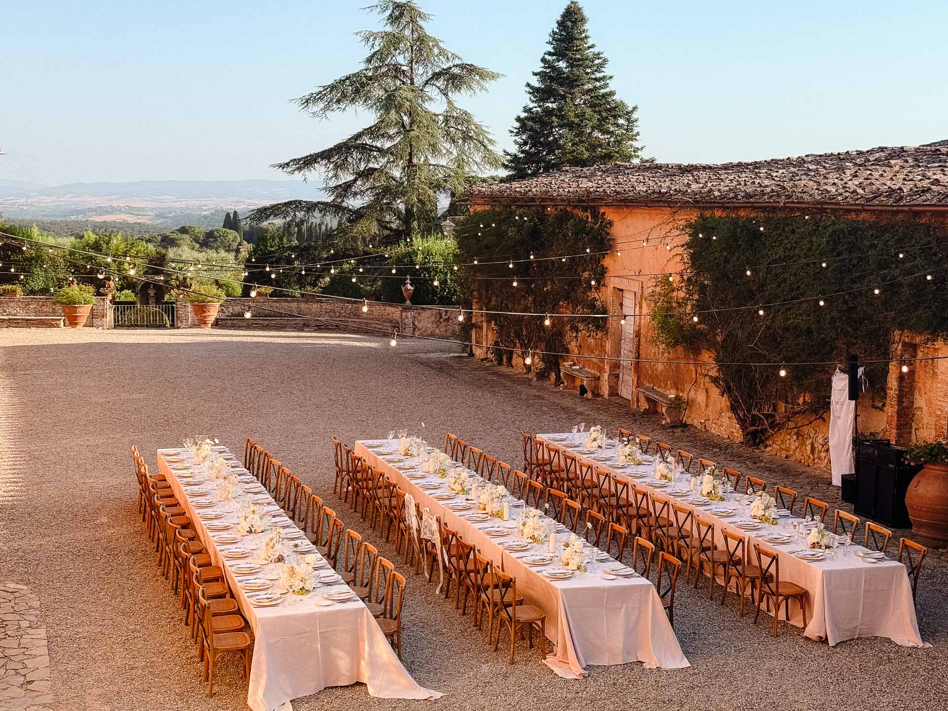 Outdoor wedding reception setup with long banquet tables, pink tablecloths, flowers, and string lights in a stone courtyard at sunset.
