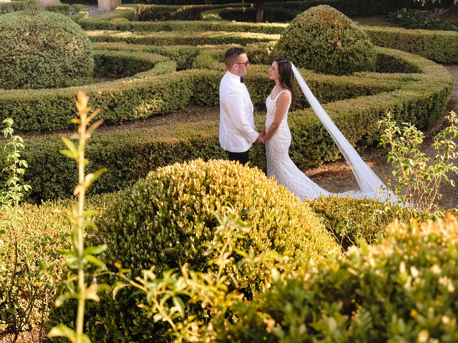 Bride and groom hold hands in a formal garden with manicured hedges; she wears a lace wedding gown and veil, he in a white suit.