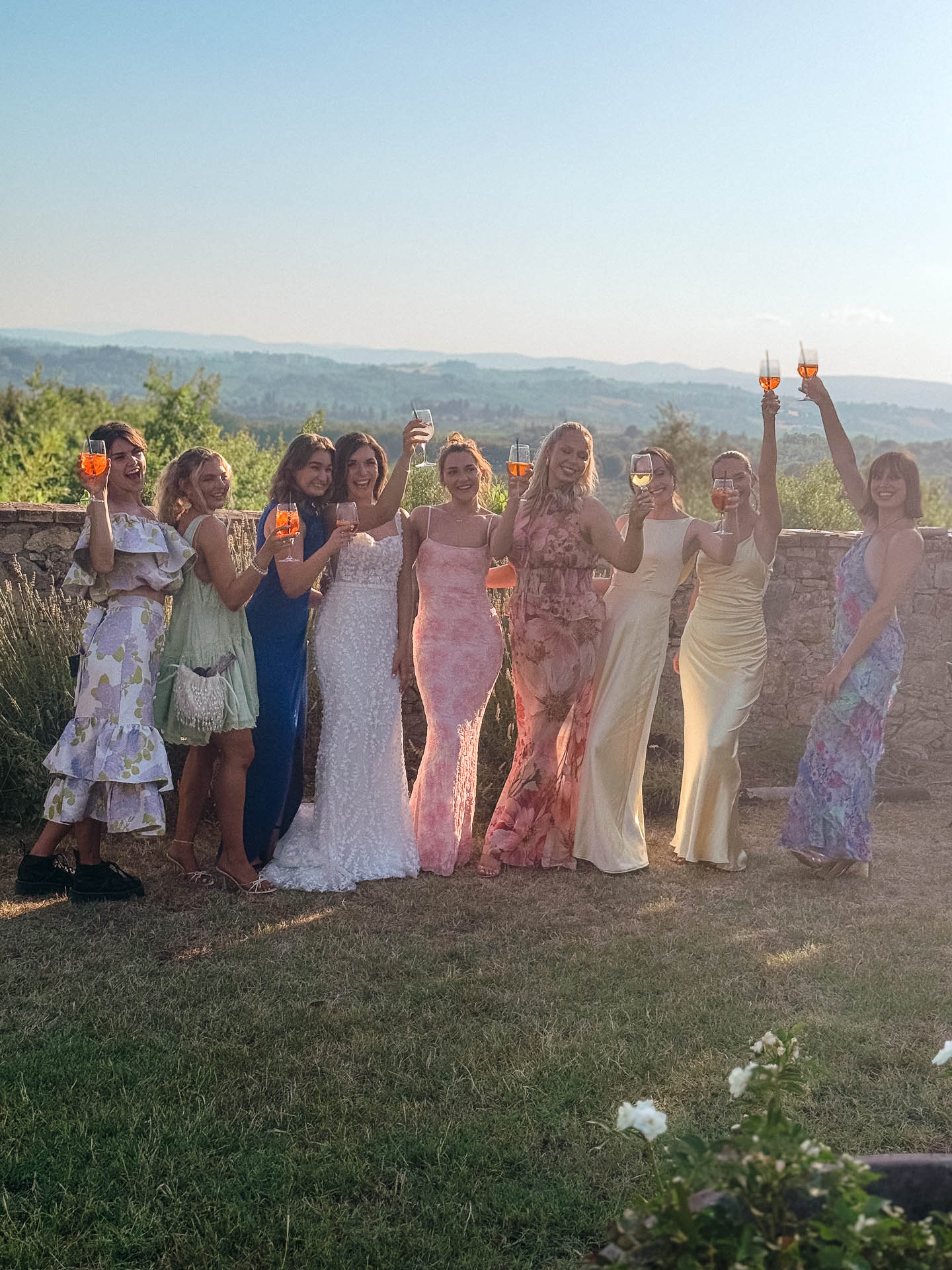 Group of women in pastel dresses raise glasses of orange wine for a toast on a sunny outdoor terrace with hills in the background.
