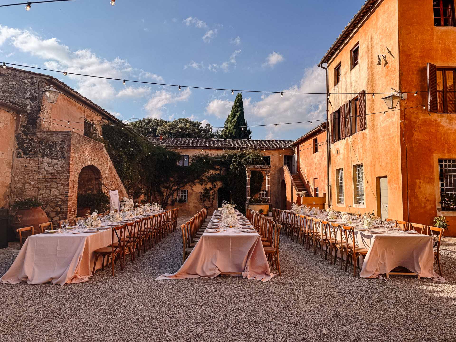 Outdoor courtyard banquet setup with long rows of tables, pink tablecloths, and string lights above the gravel yard.