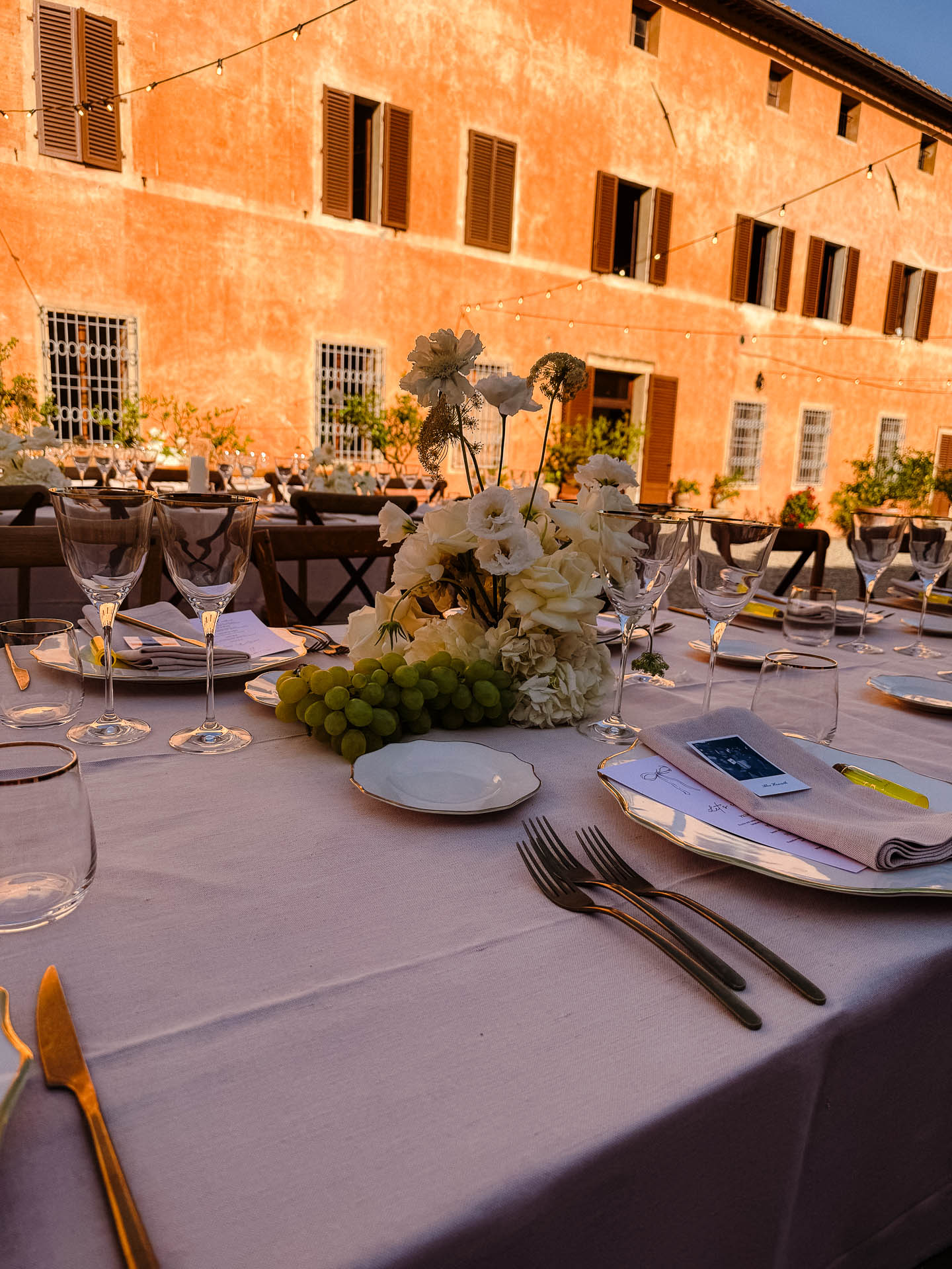 Outdoor dining table set with glassware, plates, and a floral centerpiece in a sunlit orange courtyard.
