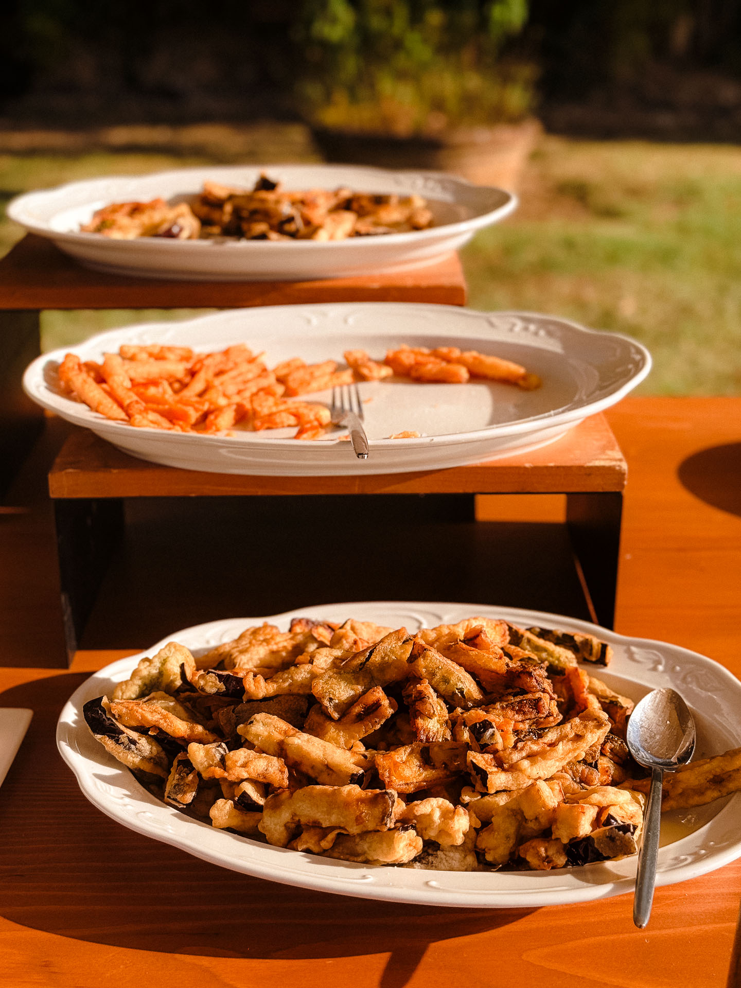 Plate of fried snack pieces on a white dish in foreground, with more plates of food on a rack in the background outdoors at dusk