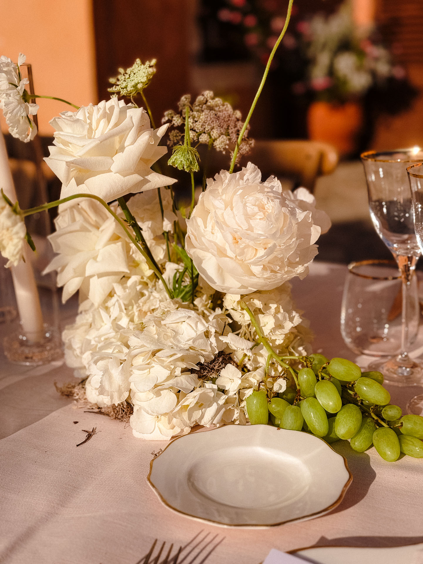 White floral centerpiece with large white roses and greenery on an elegant table setting, beside grape clusters and fine china.