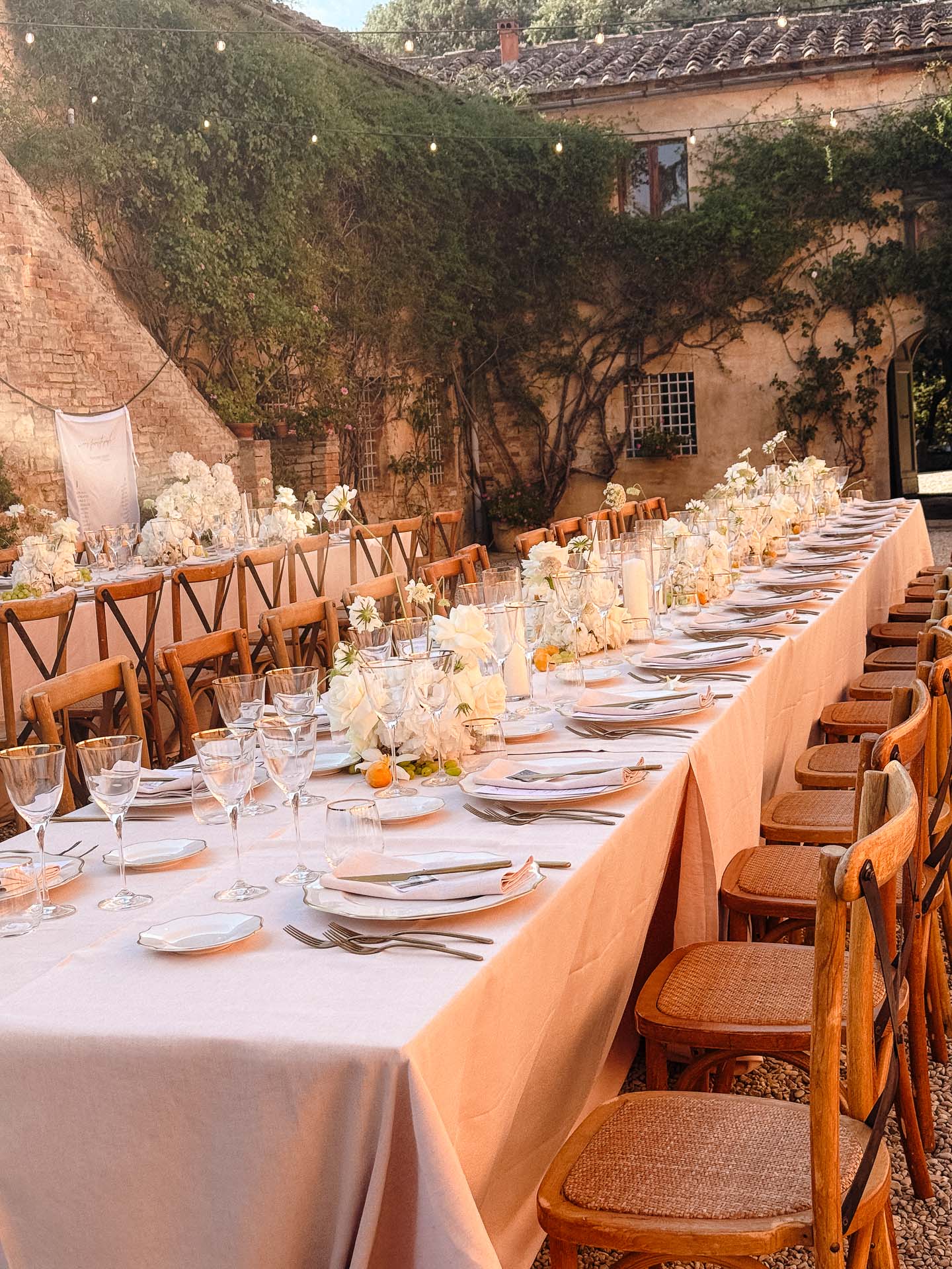 Long outdoor dining table in a stone courtyard, with white linens, flowers, and glassware under string lights.