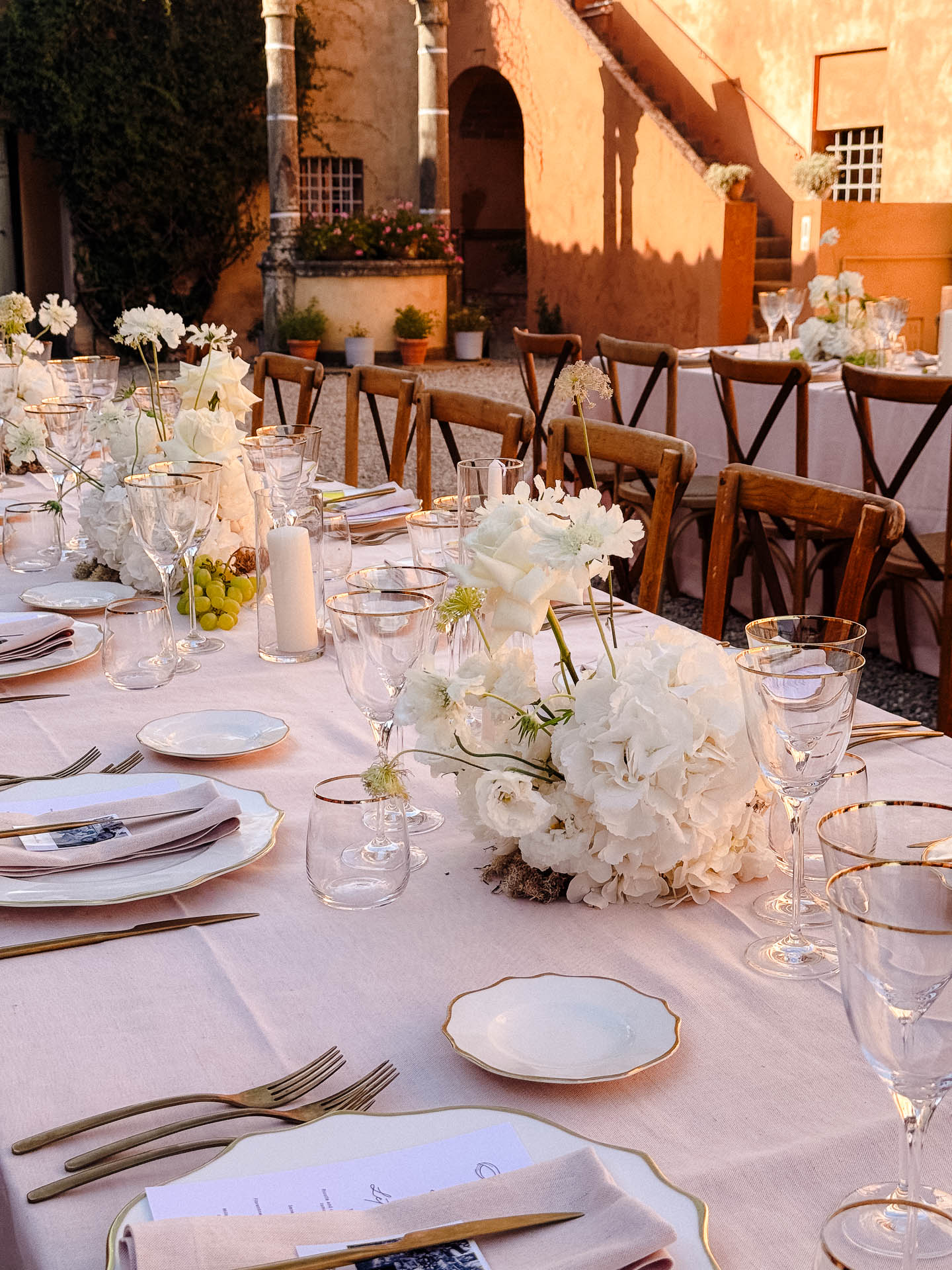 Elegant outdoor dining table set with white flowers, gold-edged plates, and glassware in a warm courtyard setting.