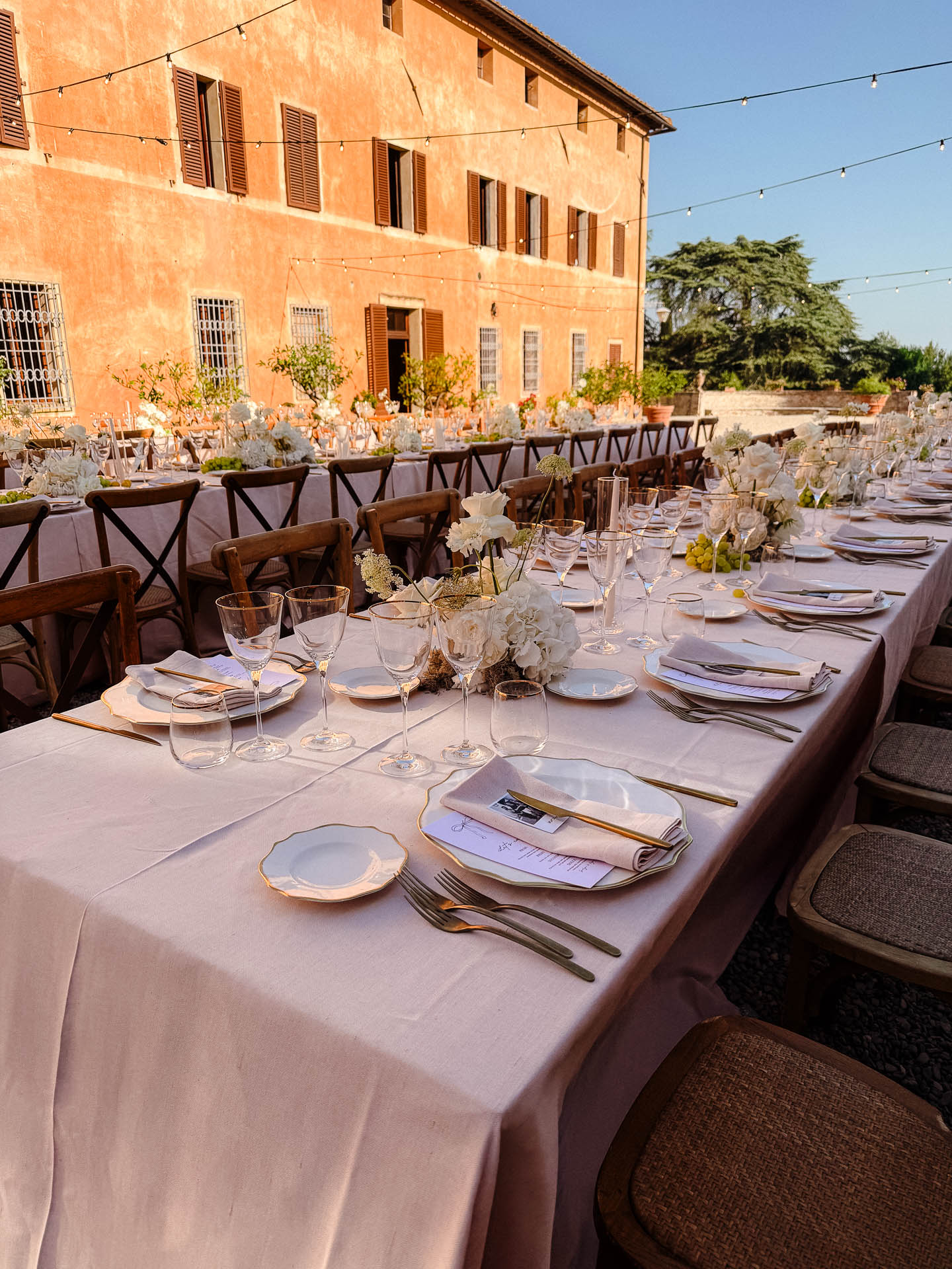 Outdoor wedding reception setup with a long white-tablecloth table, floral centerpieces, glassware, and an orange building backdrop under string lights?