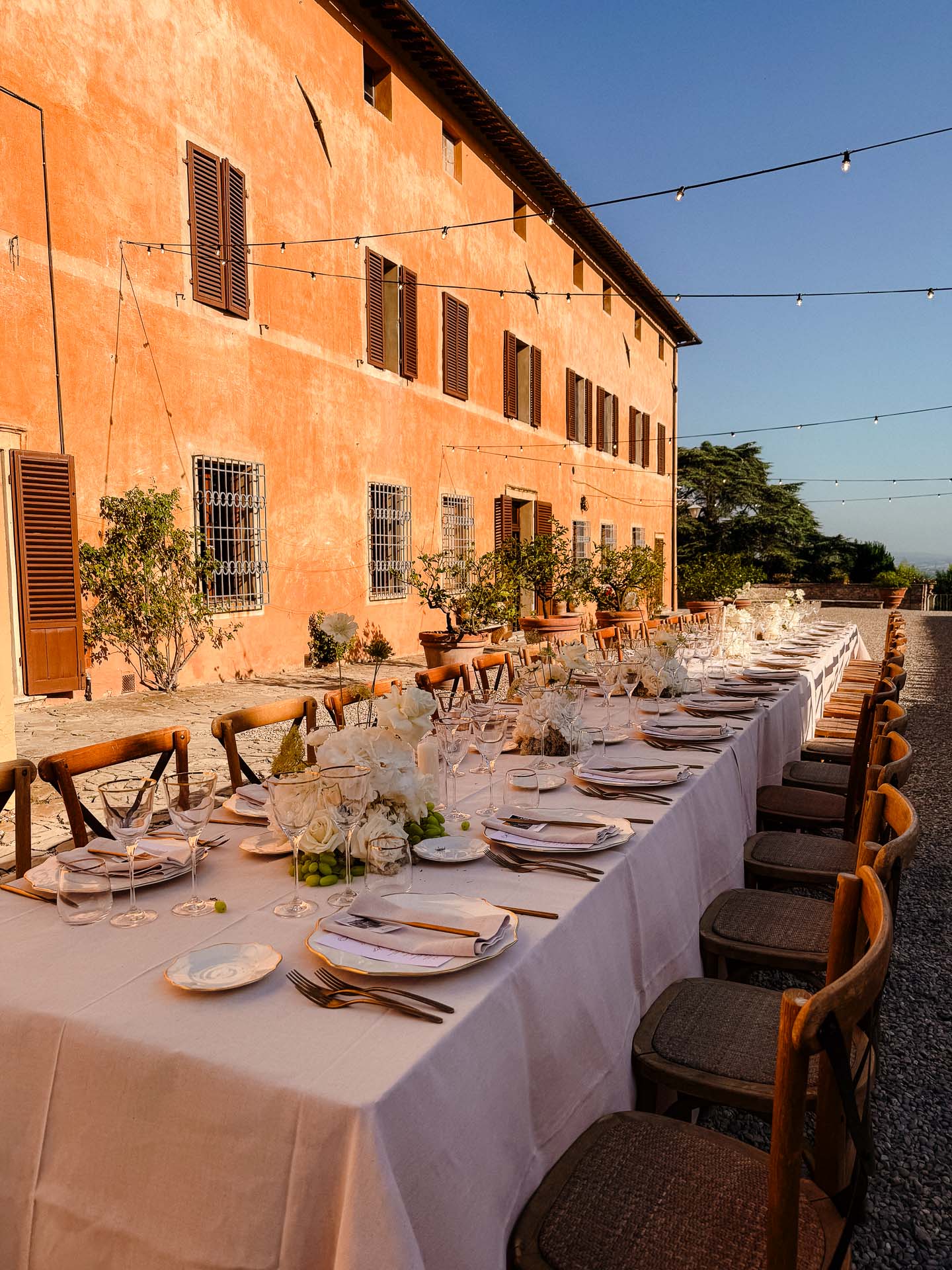 Long outdoor dining table set with white tablecloths, flowers, and glassware beside an orange stucco building under string lights at dusk.
