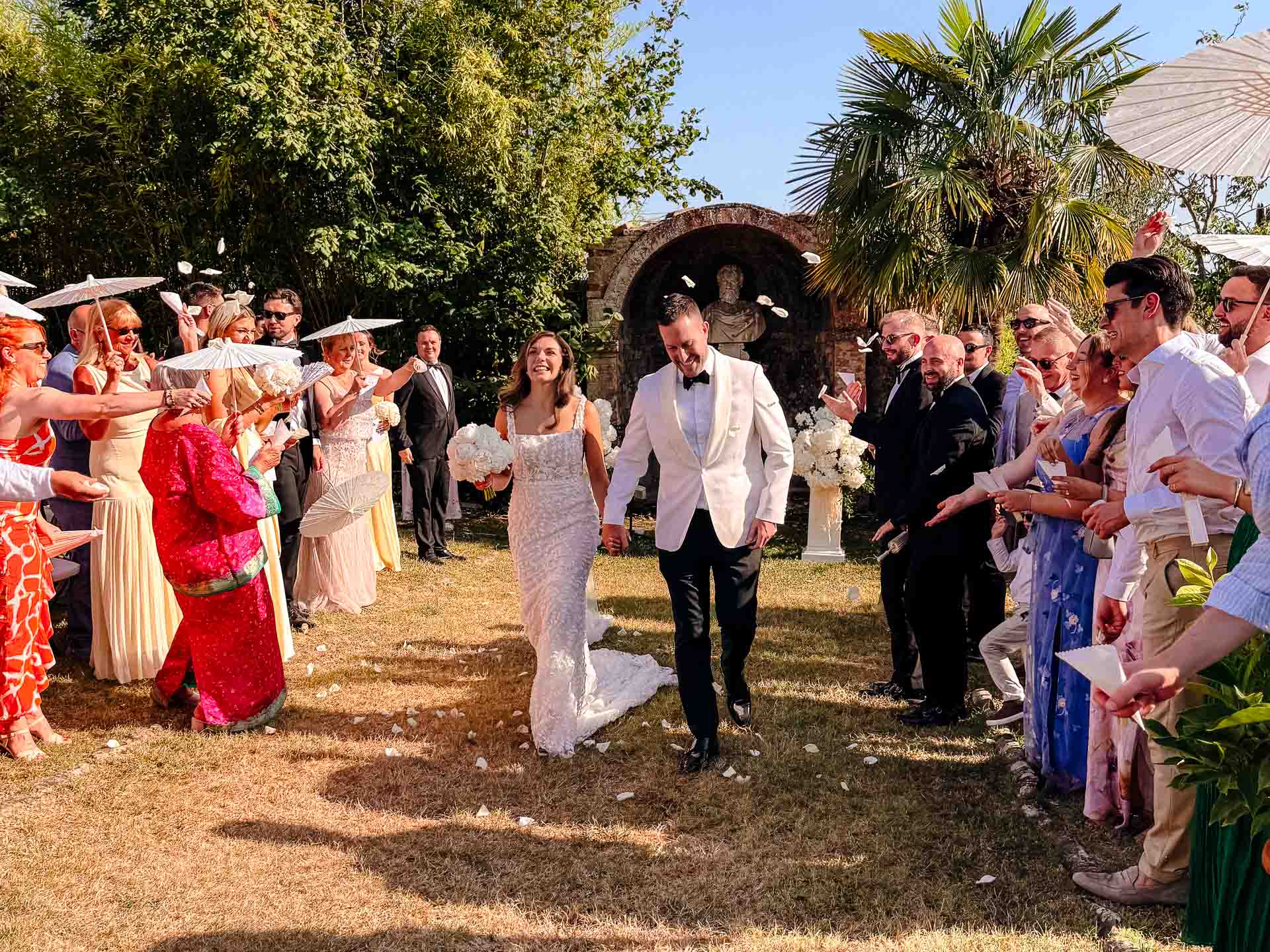 Bride and groom walk down a petal-strewn aisle outdoors as guests throw flower petals from both sides under a sunny sky.