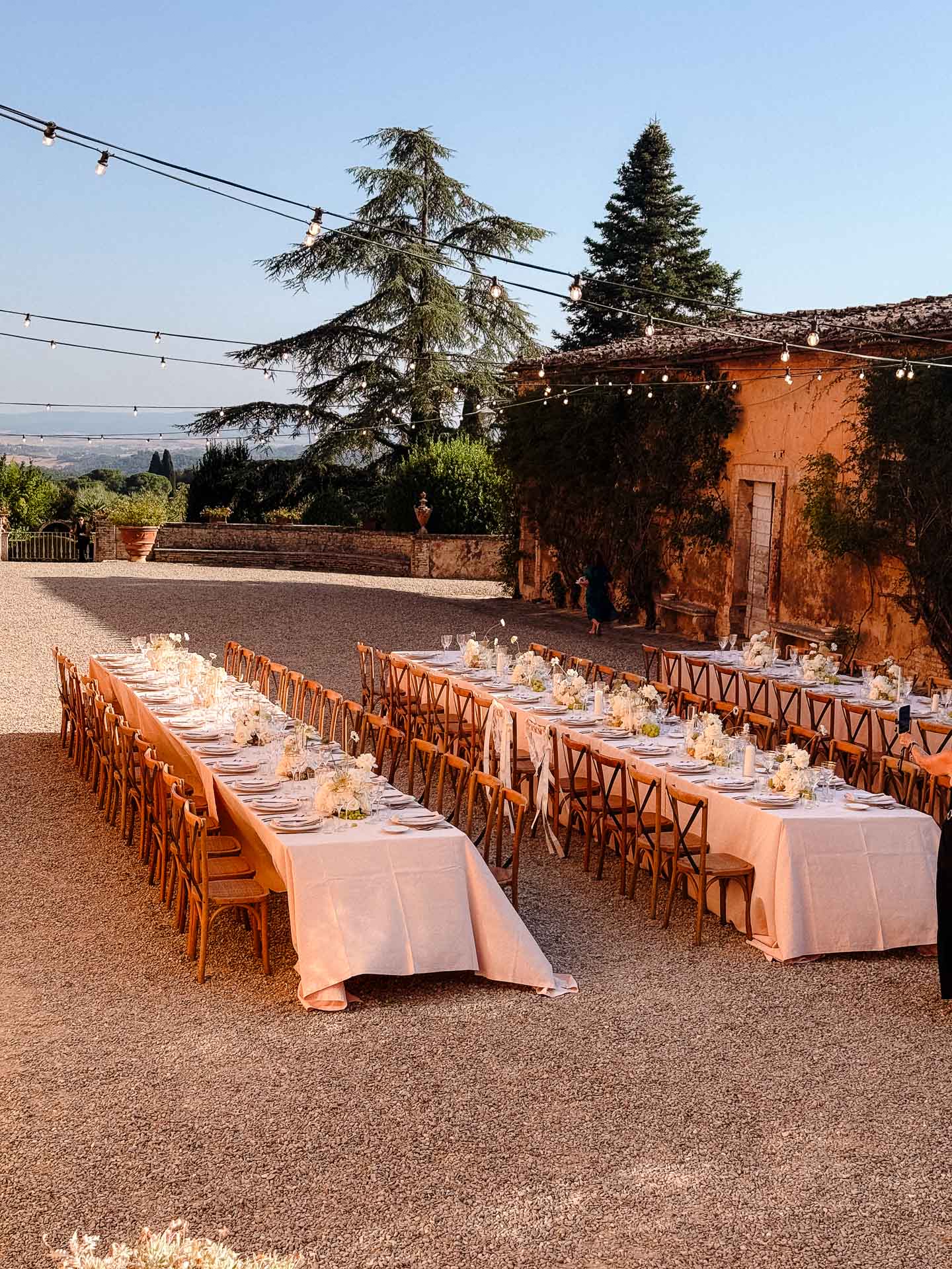Outdoor wedding or event reception setup in a sunlit stone courtyard with long rows of tables, white tablecloths, and string lights overhead.