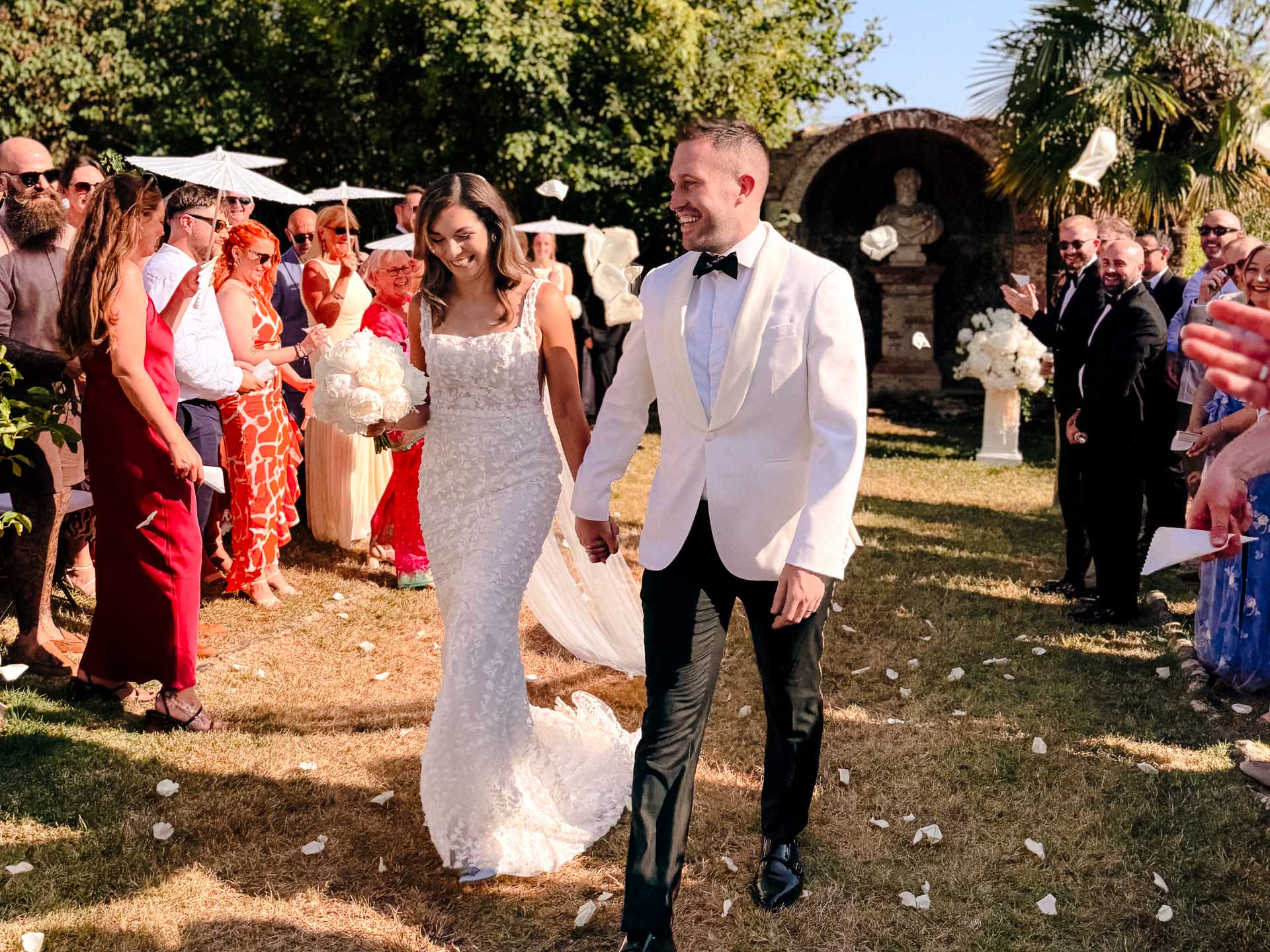 Bride in a lace wedding gown and groom in a white jacket walk hand in hand as guests cheer outdoors.