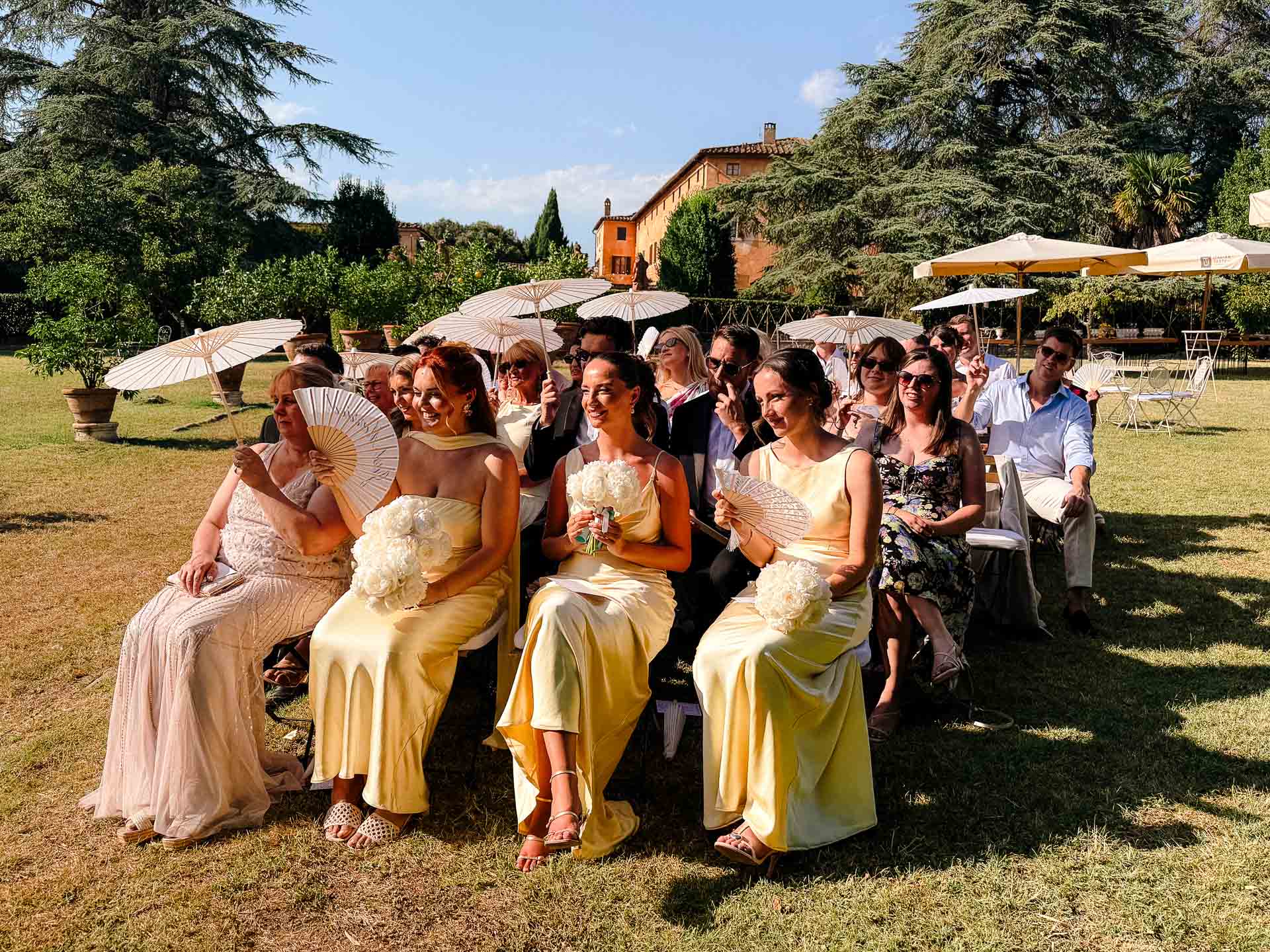 Group of wedding guests seated outdoors in bright dresses, holding white fans.