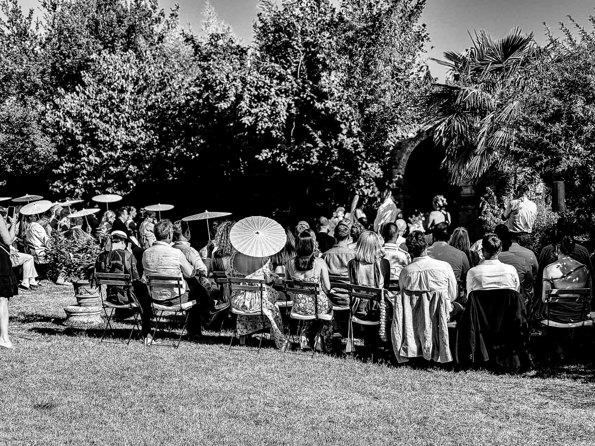 Outdoor ceremony scene with a large crowd seated on chairs in a garden, some guests holding white parasols under trees (B&W).