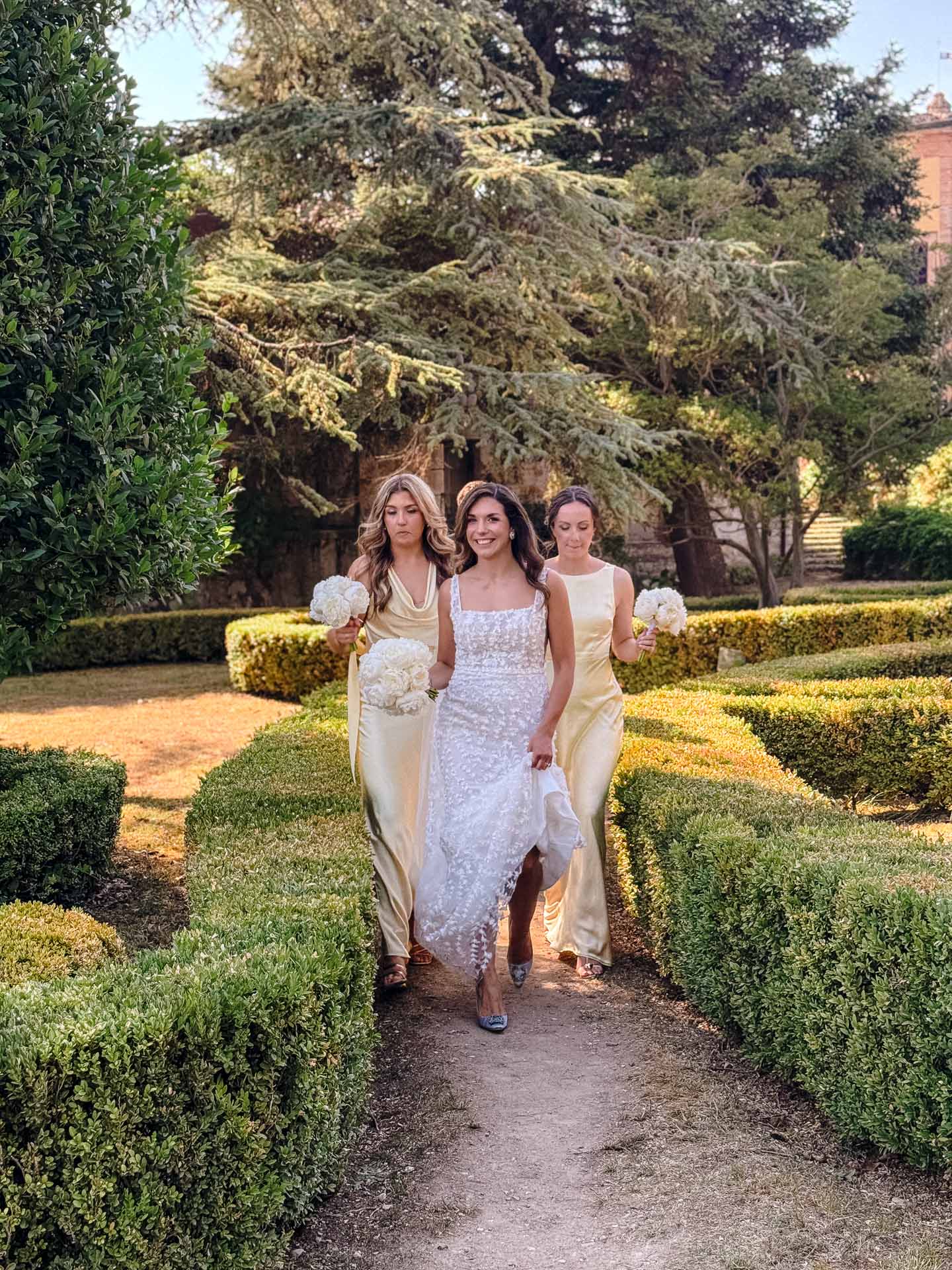 Bride in white lace gown walking with two bridesmaids in pale yellow dresses along a garden path holding white bouquets.