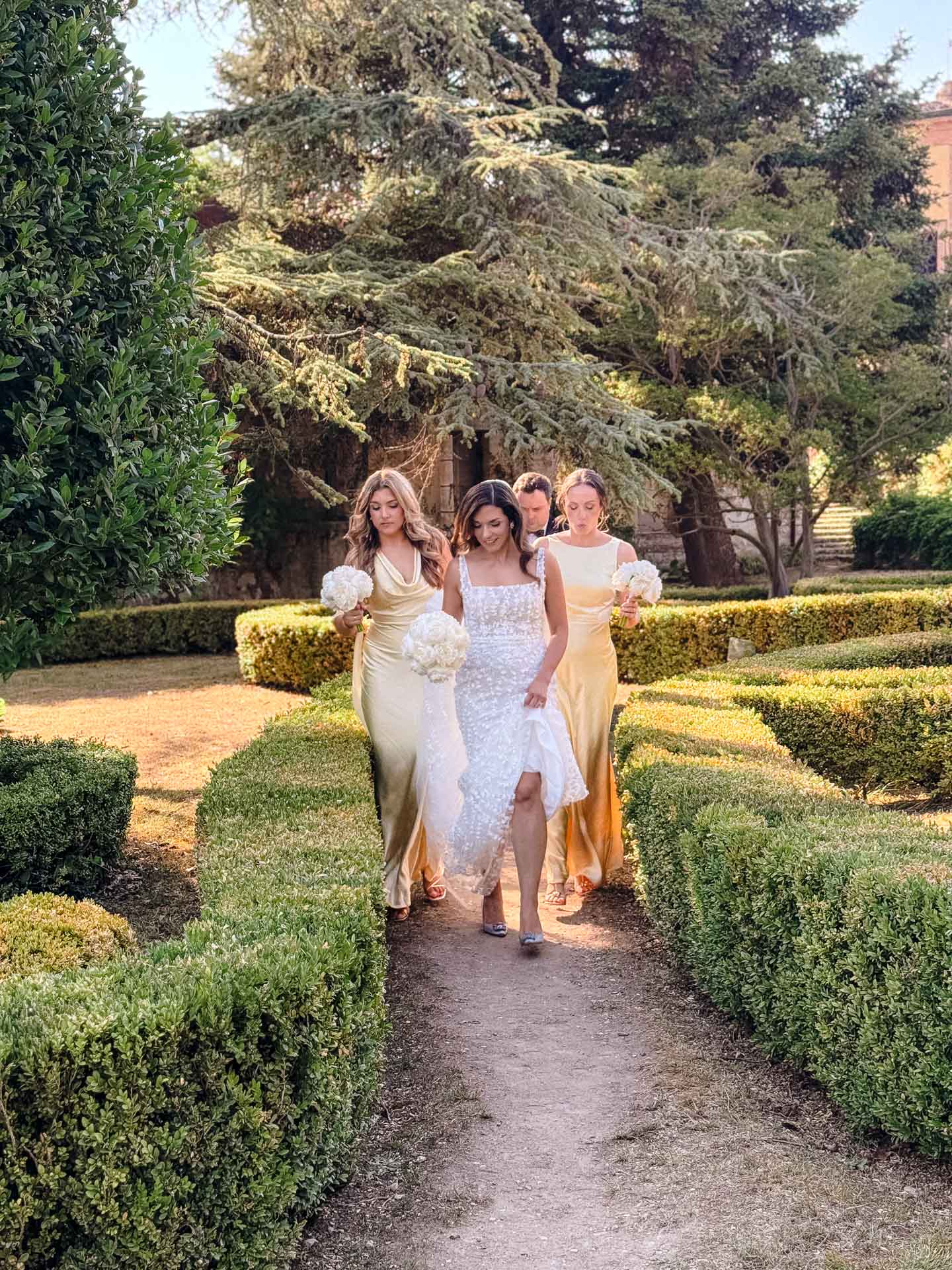 Bride in white lace gown walks with bridesmaids holding white bouquets along a garden path.