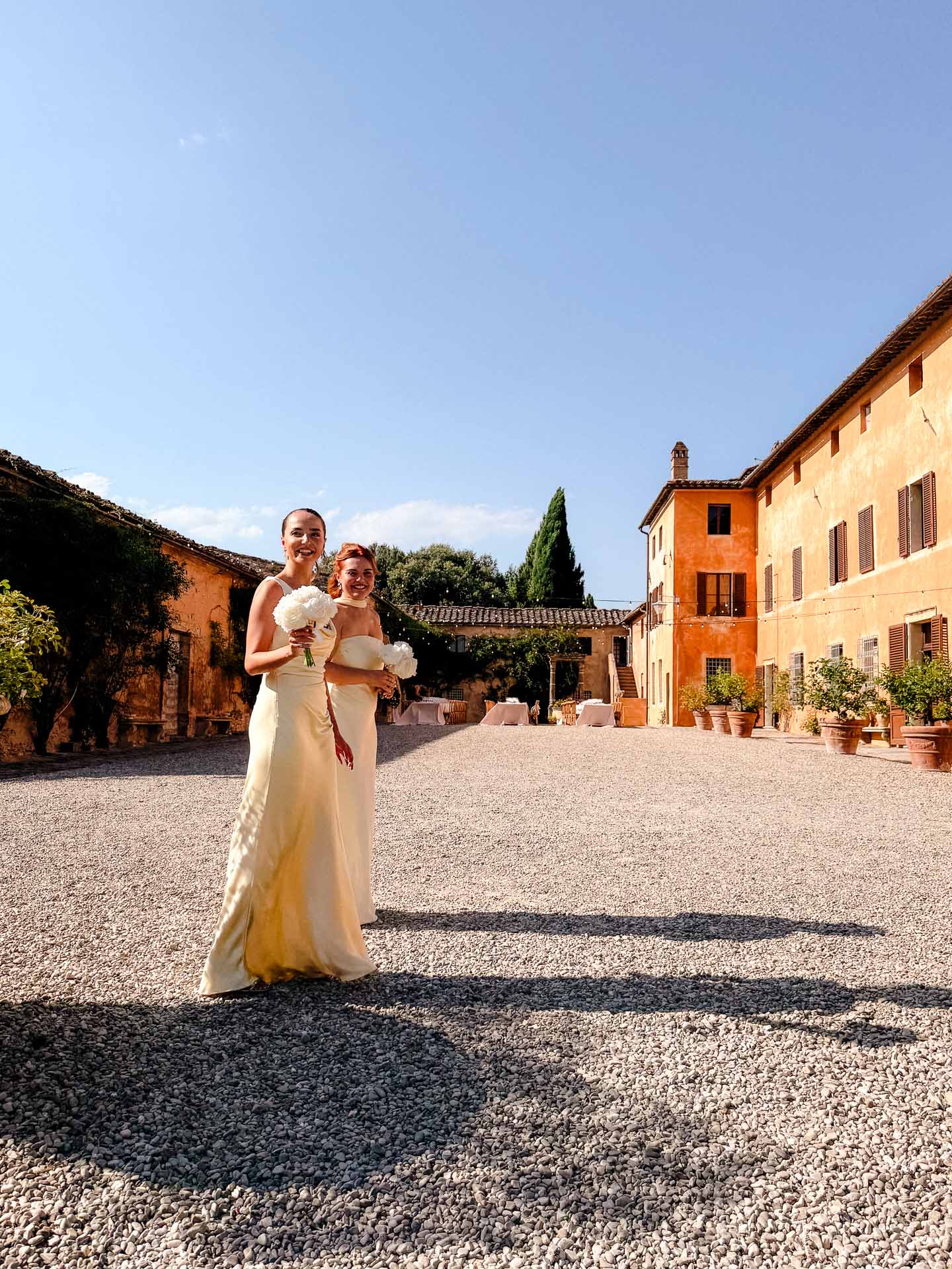 Two brides in cream gowns smile in a sunlit gravel courtyard, holding white bouquets with orange villa-style buildings in the background.