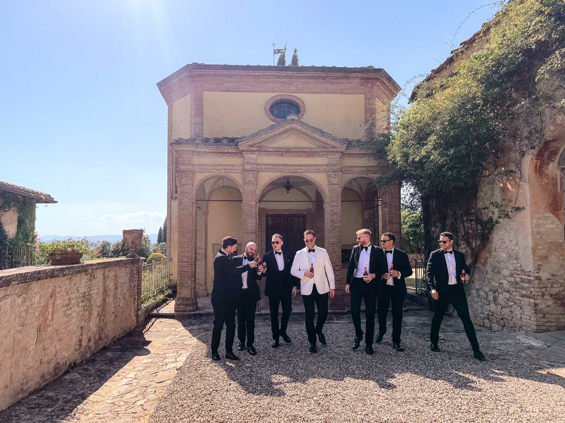 Groomsmen in tuxedos stand together in a sunlit stone courtyard in front of a historic brick building, holding beer bottles.