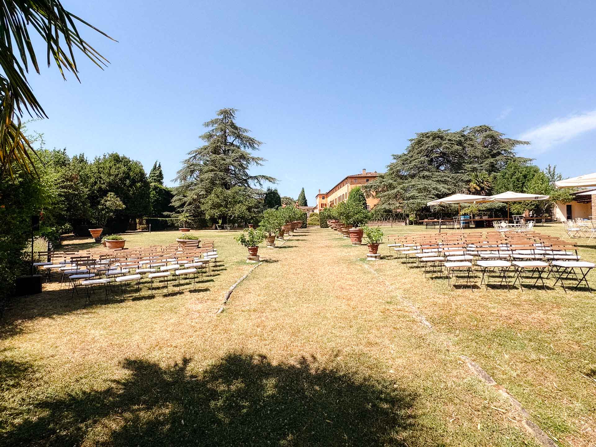Outdoor ceremony setup in a sunny garden with rows of white chairs facing a central aisle toward a brick building.