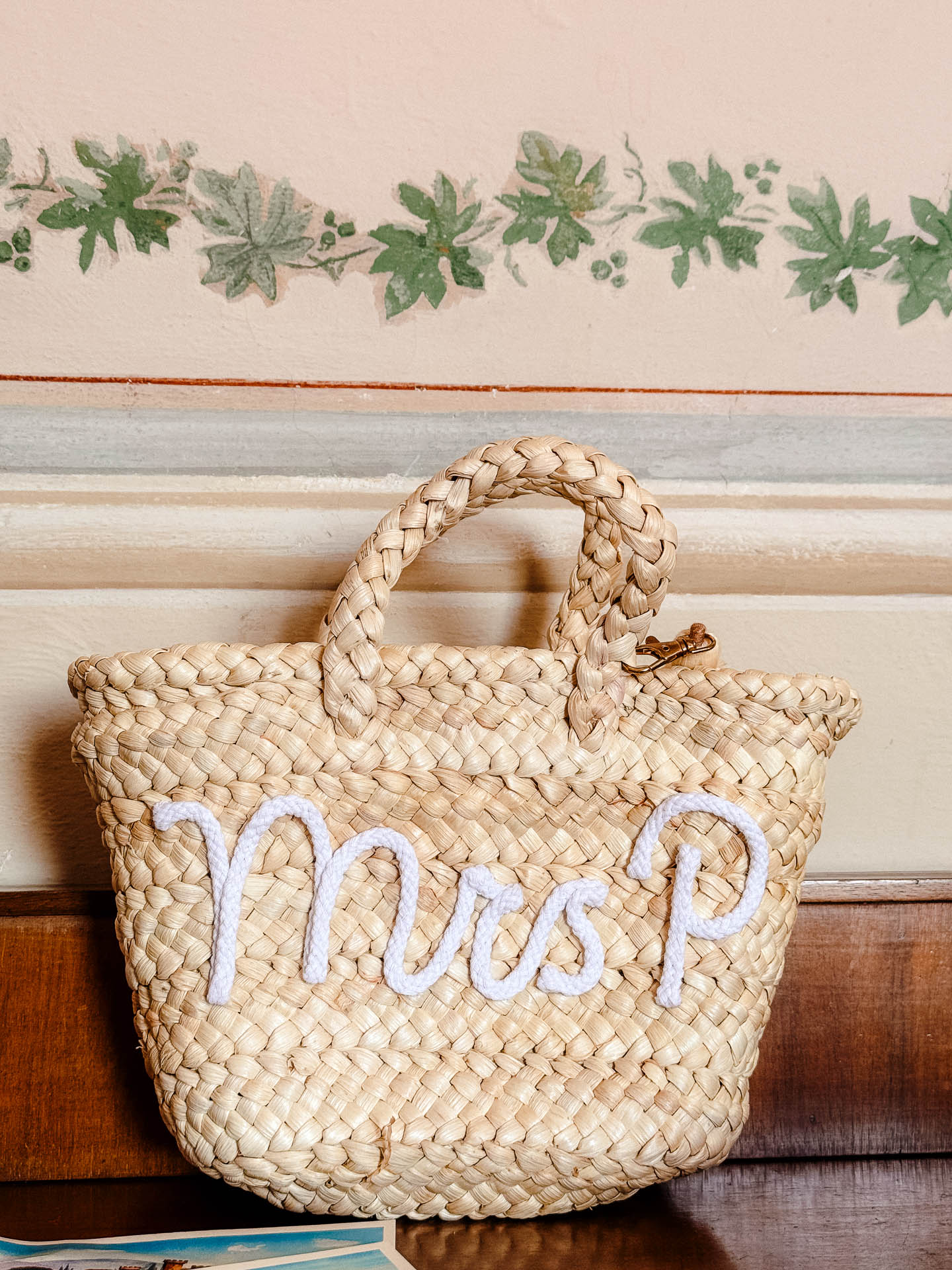Woven straw tote bag with white rope handles resting on a wooden surface against a pastel wall with green vine decor.