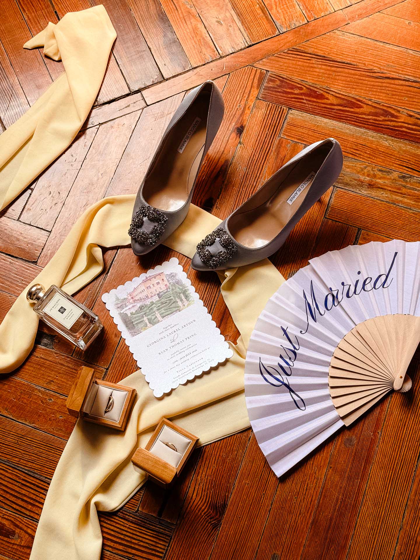 Gray high heels with jeweled buckles placed on pale satin, surrounded by wedding invitation, perfume, and a 'Married' fan on a wooden floor.