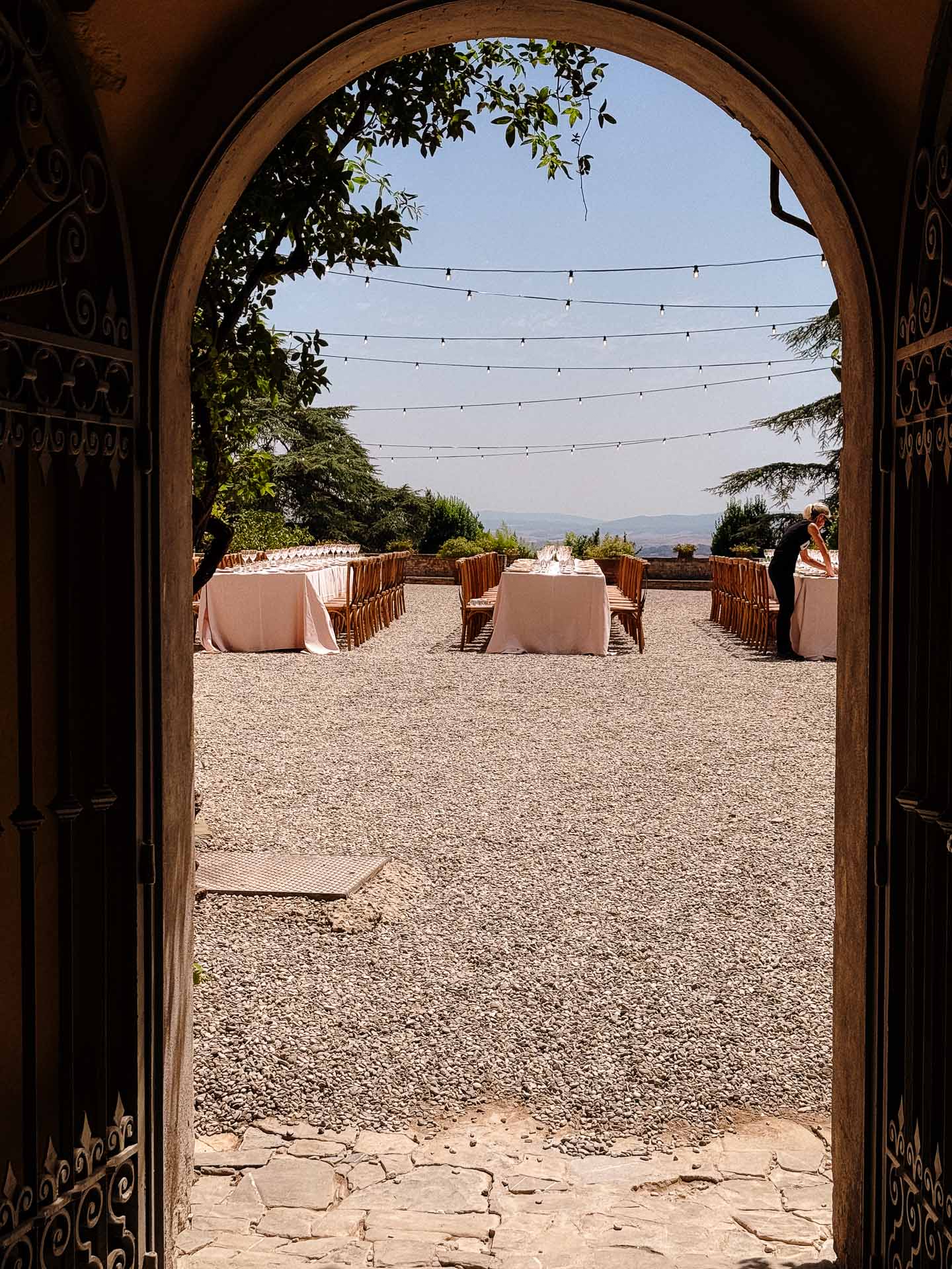 Outdoor event setup seen through an arched doorway: long tables with pink tablecloths, string lights overhead.