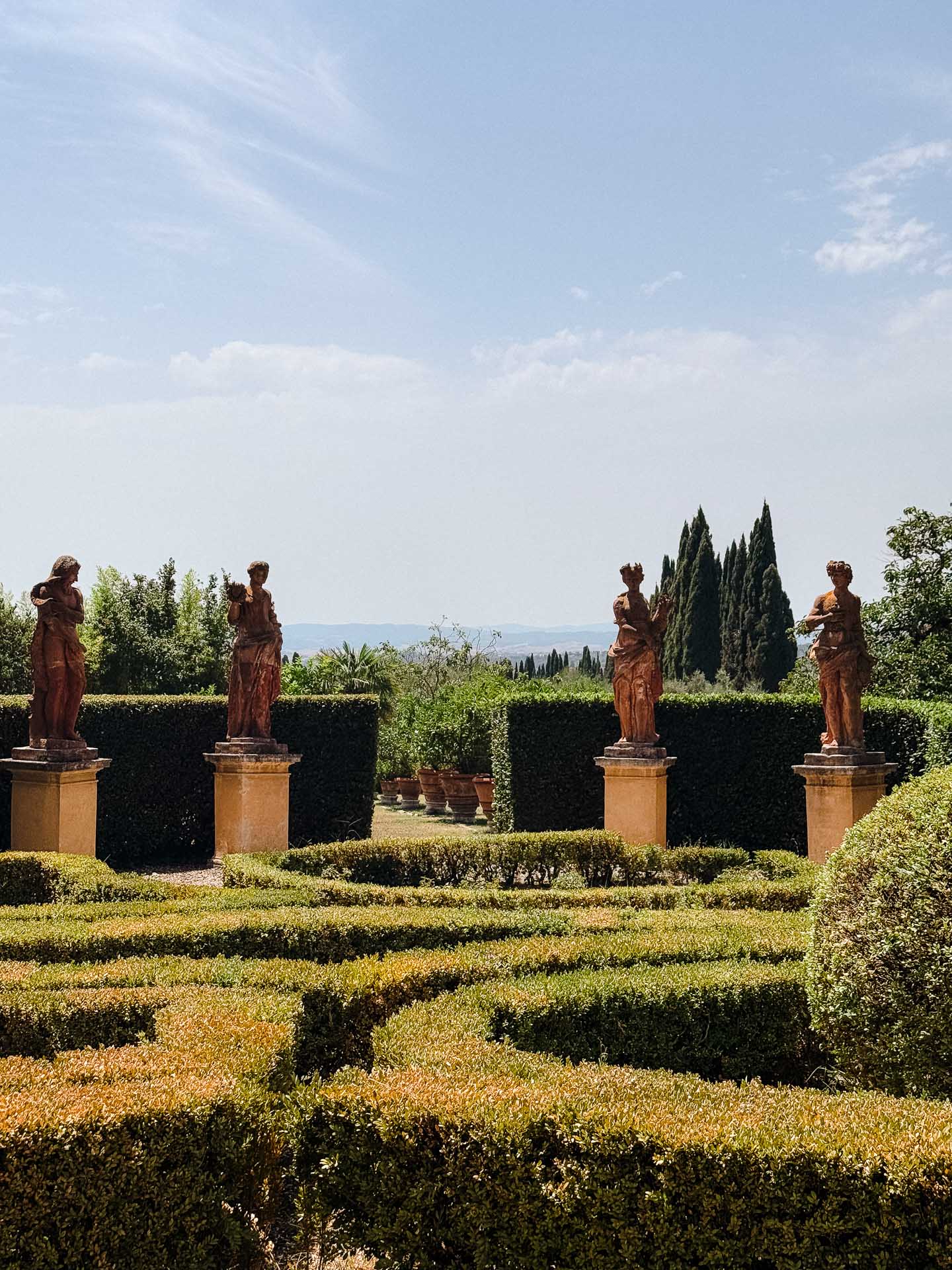 Row of classical statues on pedestals in a formal garden with manicured hedges and blue sky above.