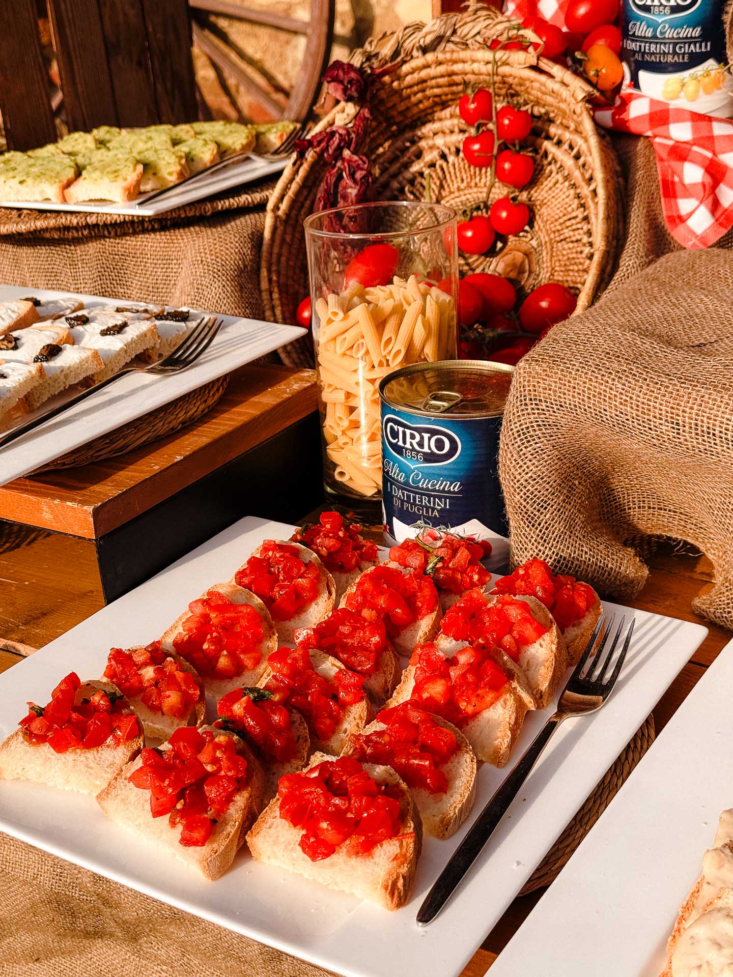 Bruschetta topped with diced tomatoes on a white platter, alongside pasta in a glass and a can of Cirio tomato product in a rustic display.